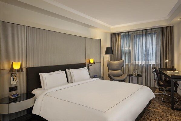 A hotel room with a large bed, two bedside lamps, a desk with a chair, and a view of the outside through curtains.