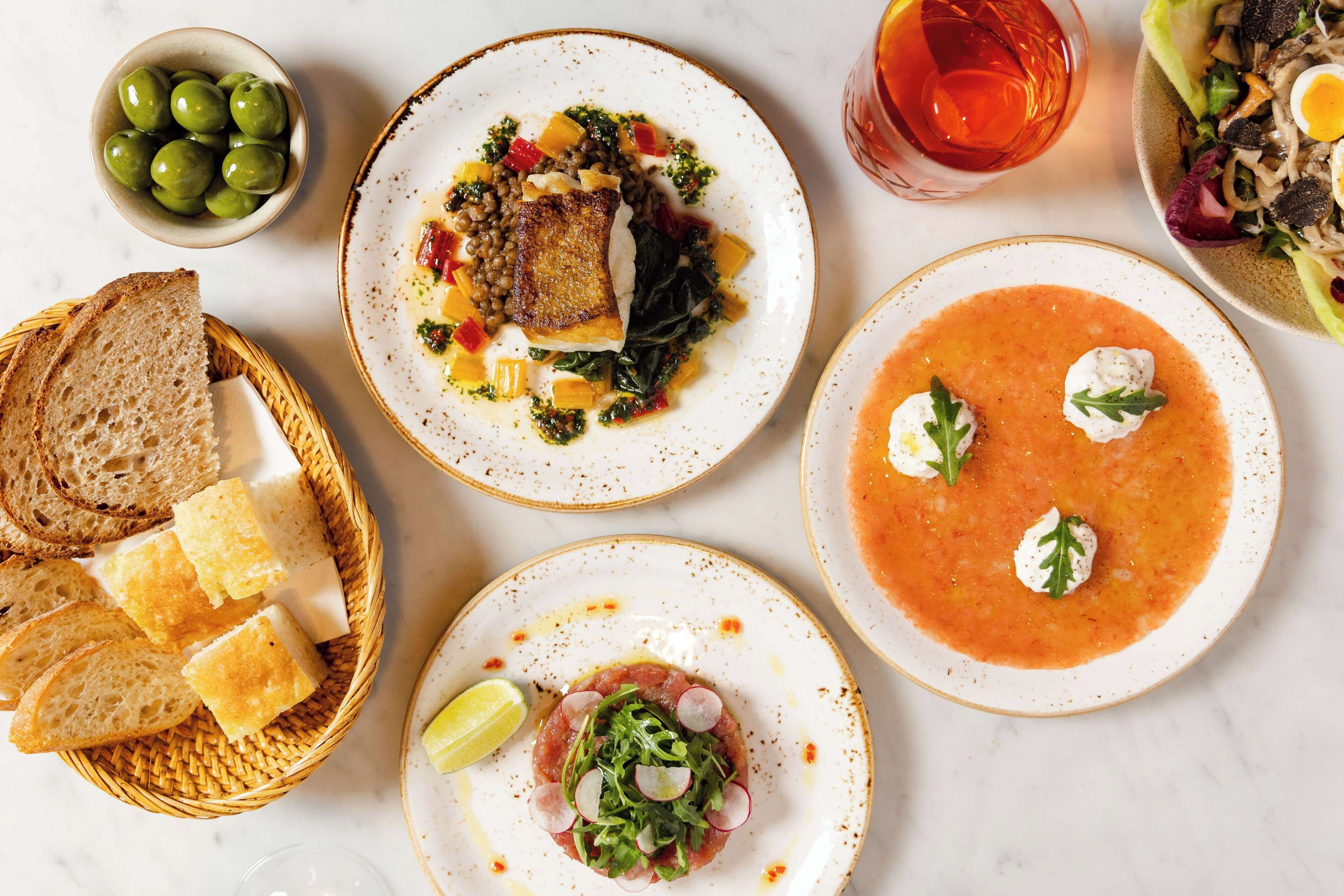 A variety of dishes including a plate with a fish fillet, a bowl of soup, and a salad. There are also slices of bread and a small bowl of olives.