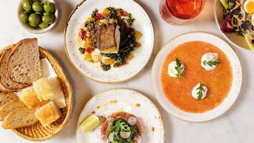 A variety of dishes including a plate with a fish fillet, a bowl of soup, and a salad. There are also slices of bread and a small bowl of olives.