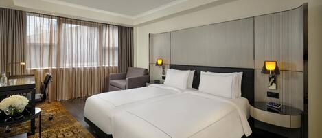 Habitación de hotel con una cama grande, un escritorio con silla, una mesita con un jarrón de flores y vistas a la ciudad a través de amplios ventanales.