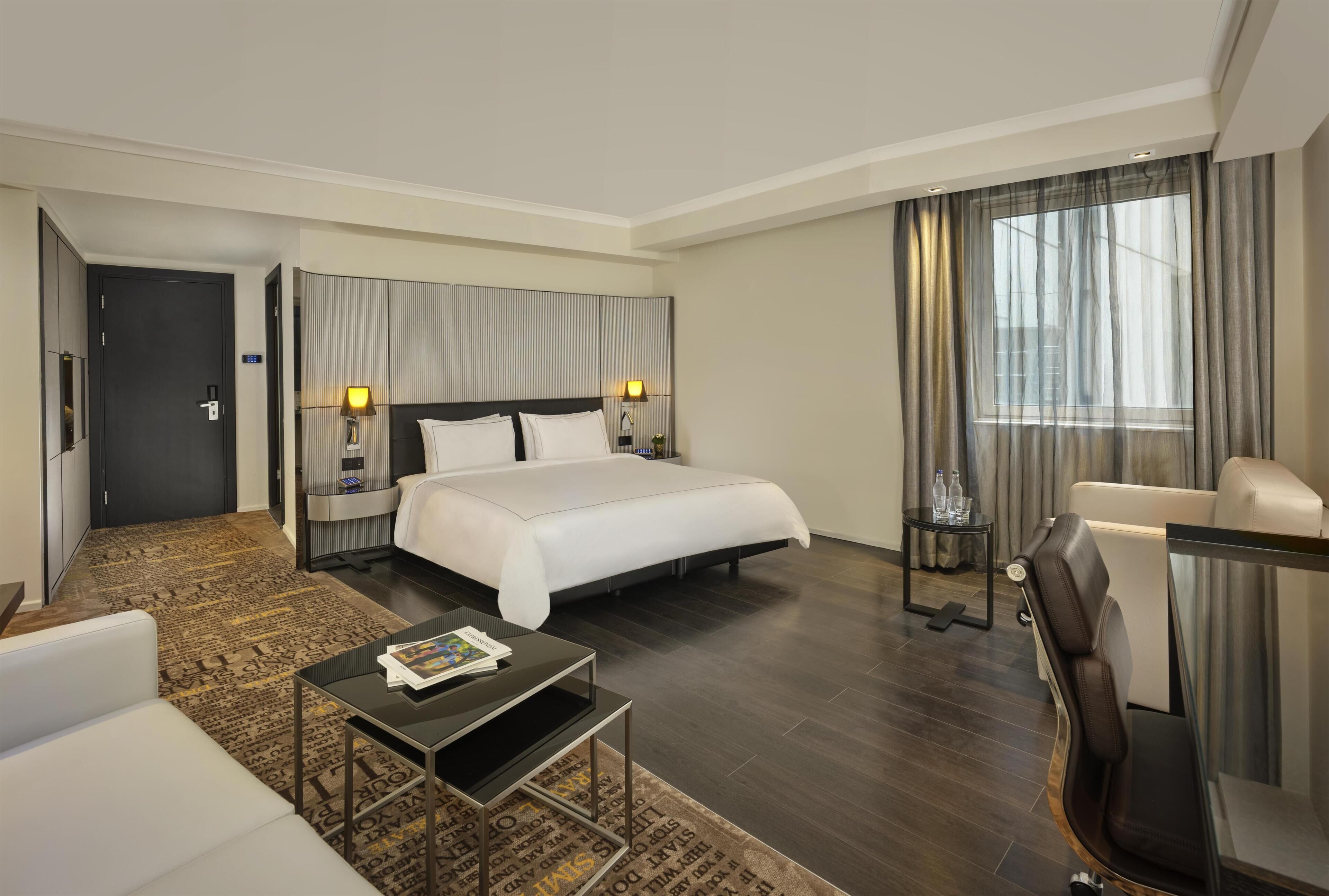 Ein Hotelzimmer mit Bett, Schreibtisch, Stuhl, kleinem Tisch und einem Fenster mit Vorhängen.