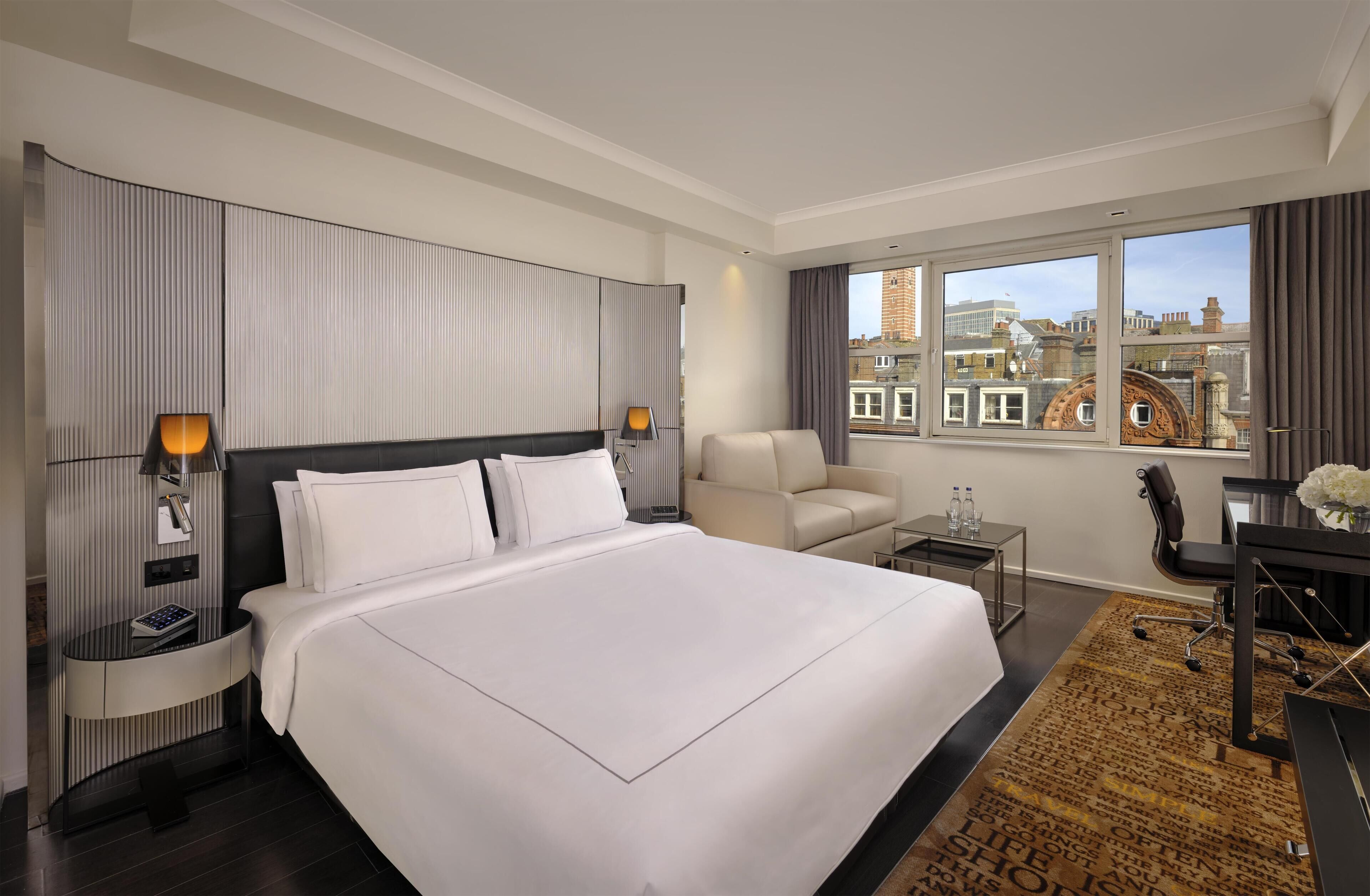 Une chambre d’hôtel moderne, dotée d’un grand lit, d’un bureau, d’une chaise, d’une petite table et offrant une vue sur la ville grâce à de grandes fenêtres.