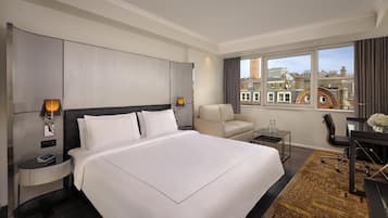 A modern hotel room with a large bed, a desk, a chair, a small table, and a view of the city through large windows.