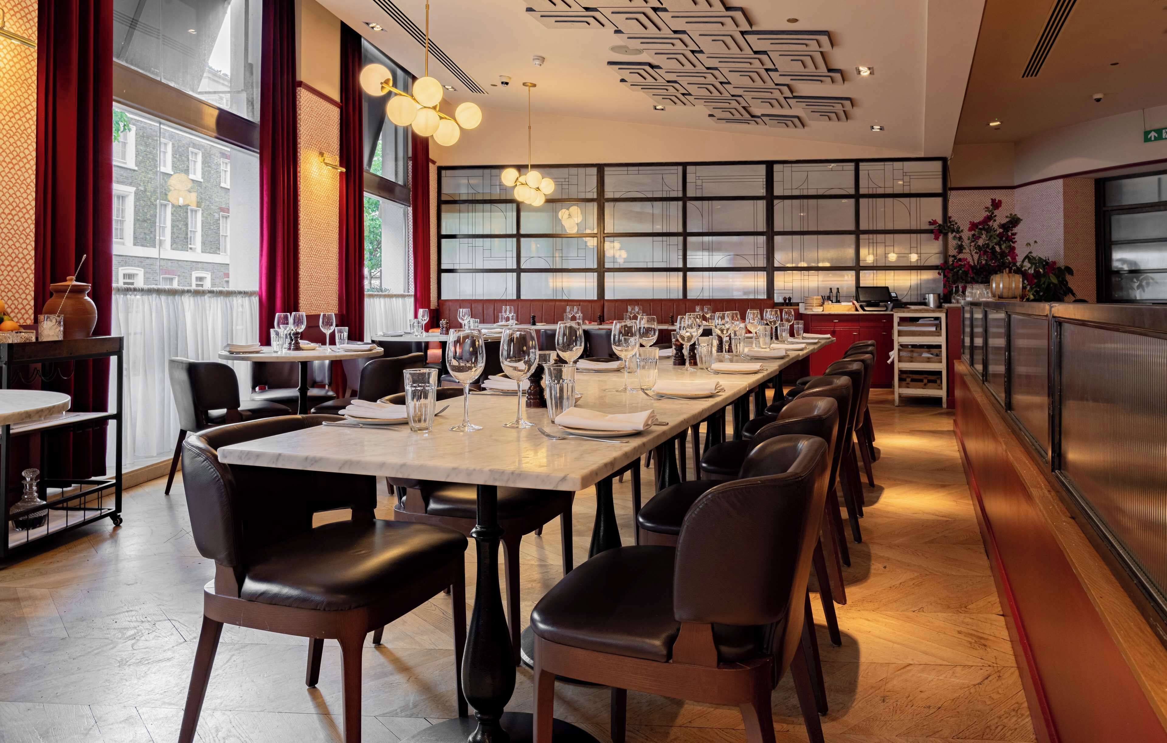 A restaurant with long tables set for dining, a bar area, and large windows with a city view.