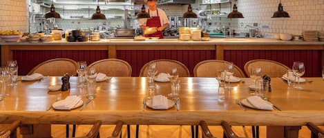 Un restaurant avec une longue table en bois dressée pour le repas, un chef qui prépare les plats au comptoir et un mur recouvert de carreaux dans la cuisine.