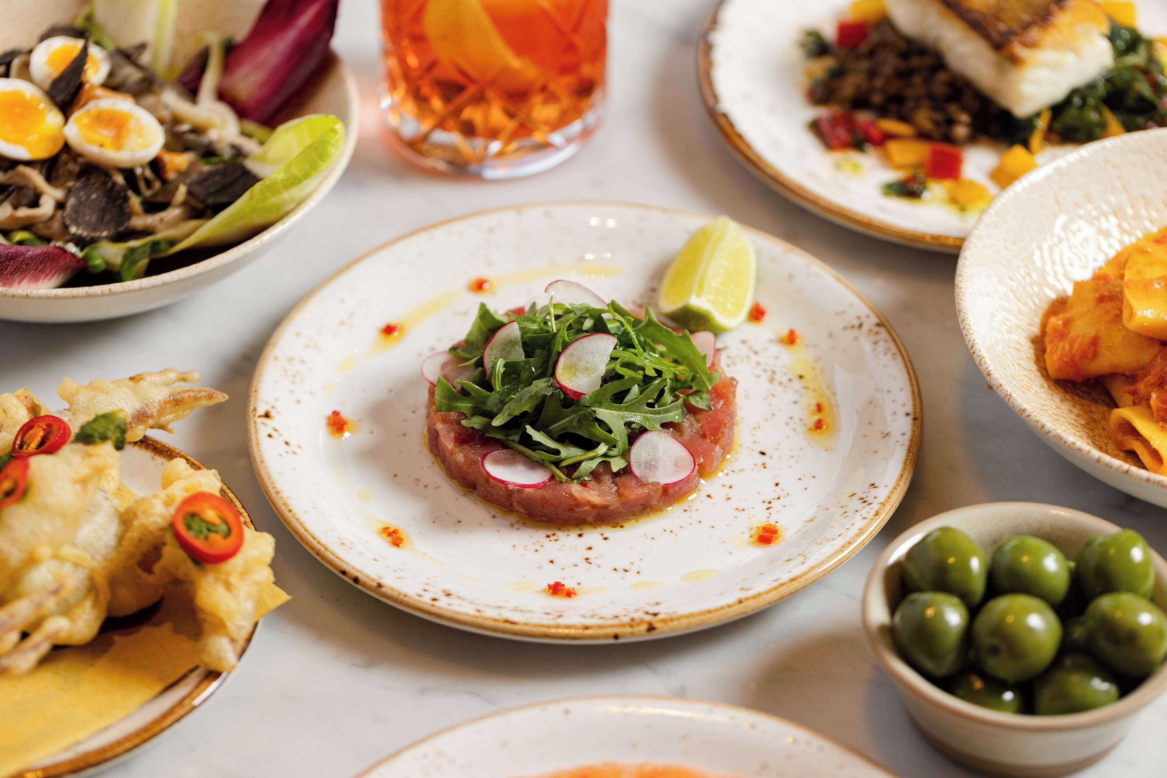 Diversos pratos em uma mesa, incluindo um prato com tartare de carne coberto com fatias de rabanete e um pedaço de limão.