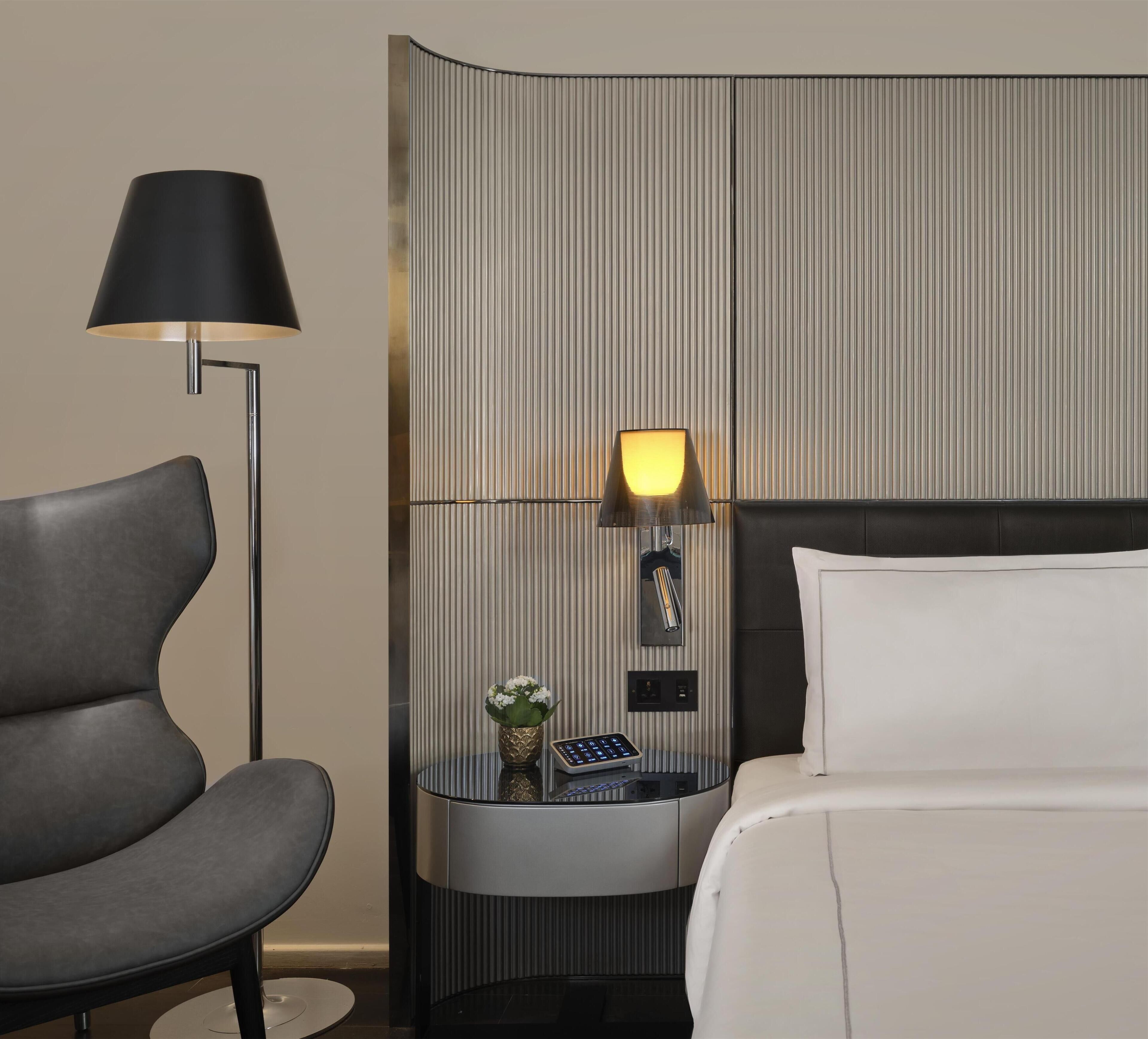 A hotel room with a bed, a chair, a nightstand with a lamp, and a control panel.