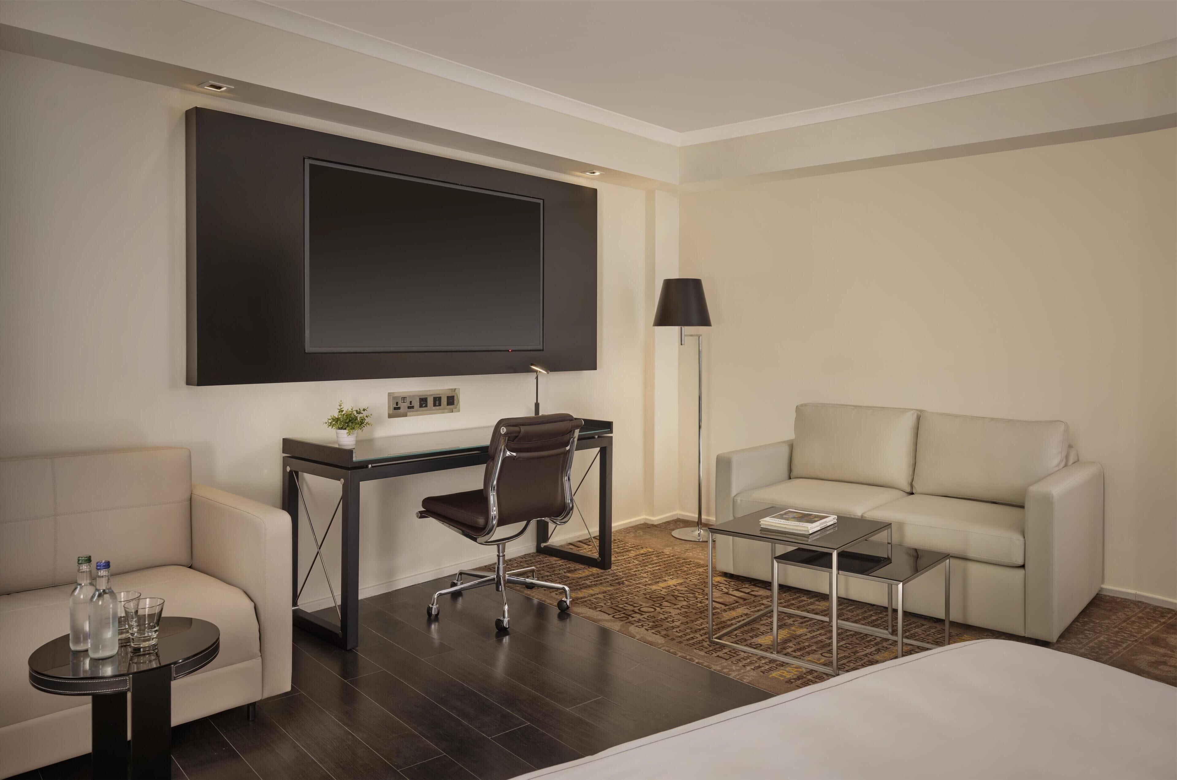 A hotel room with a bed, a desk with a chair, a sofa, a TV, and a lamp.