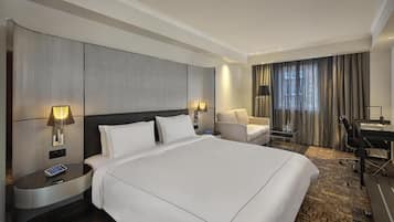 Habitación de hotel con una cama grande, dos mesitas de noche con lámparas, un escritorio, una silla y vistas a la ciudad desde la ventana.