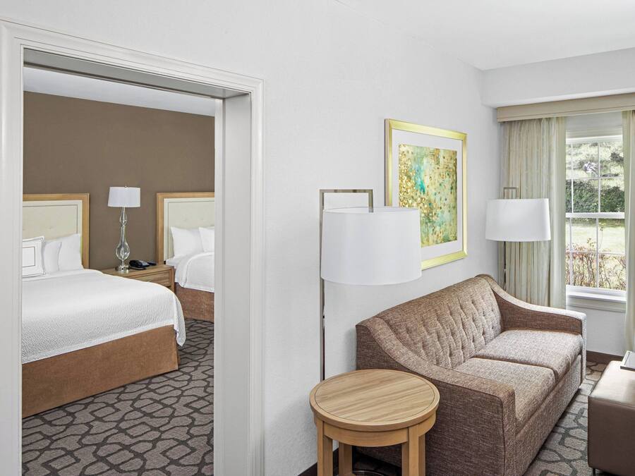 A hotel room with a bed, two bedside tables, a sofa, a round wooden stool, a lamp, and a painting on the wall.