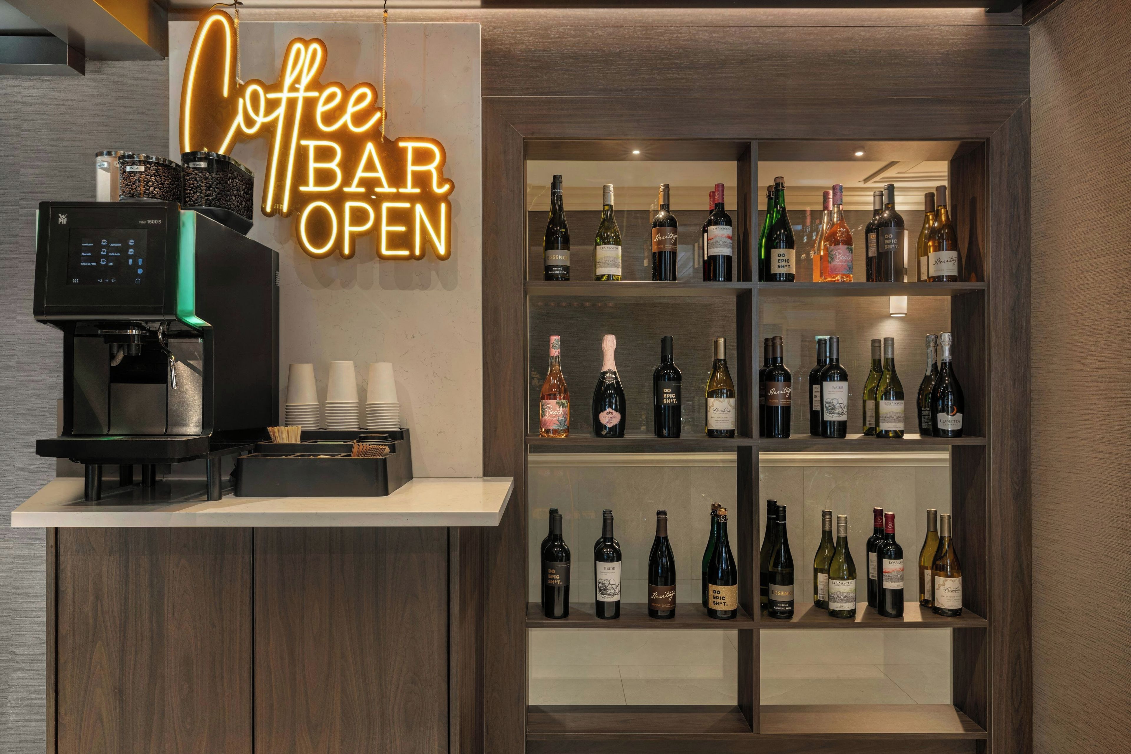 A coffee bar with a coffee machine and a neon sign that reads 'Coffee Bar Open'. There are shelves with various bottles of wine and spirits.