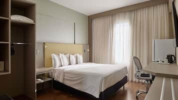 Une chambre d’hôtel comprenant un lit, un bureau avec une chaise, une armoire et une grande fenêtre avec des rideaux.