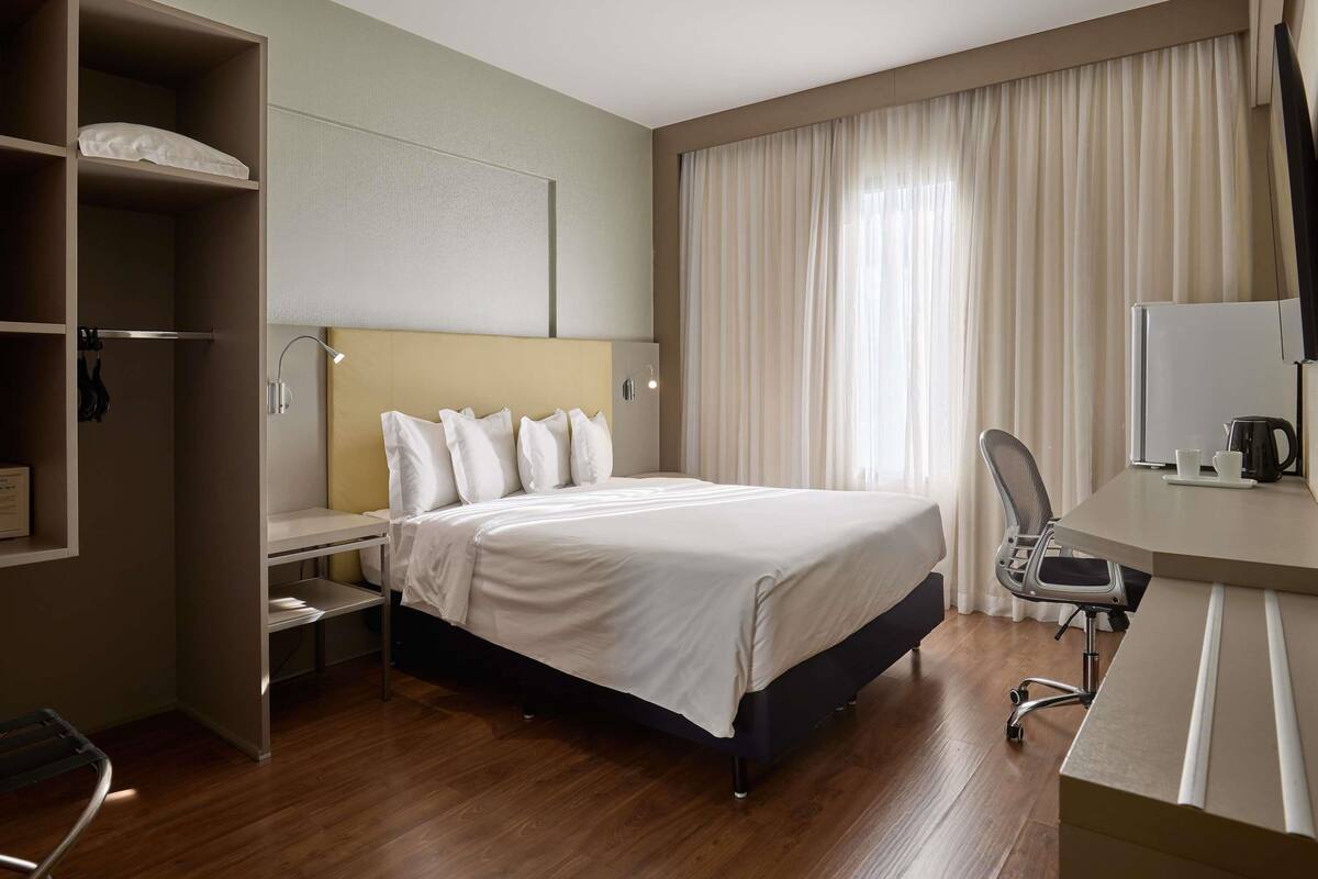 A hotel room with a bed, a desk, a chair, a wardrobe, and a window with curtains.