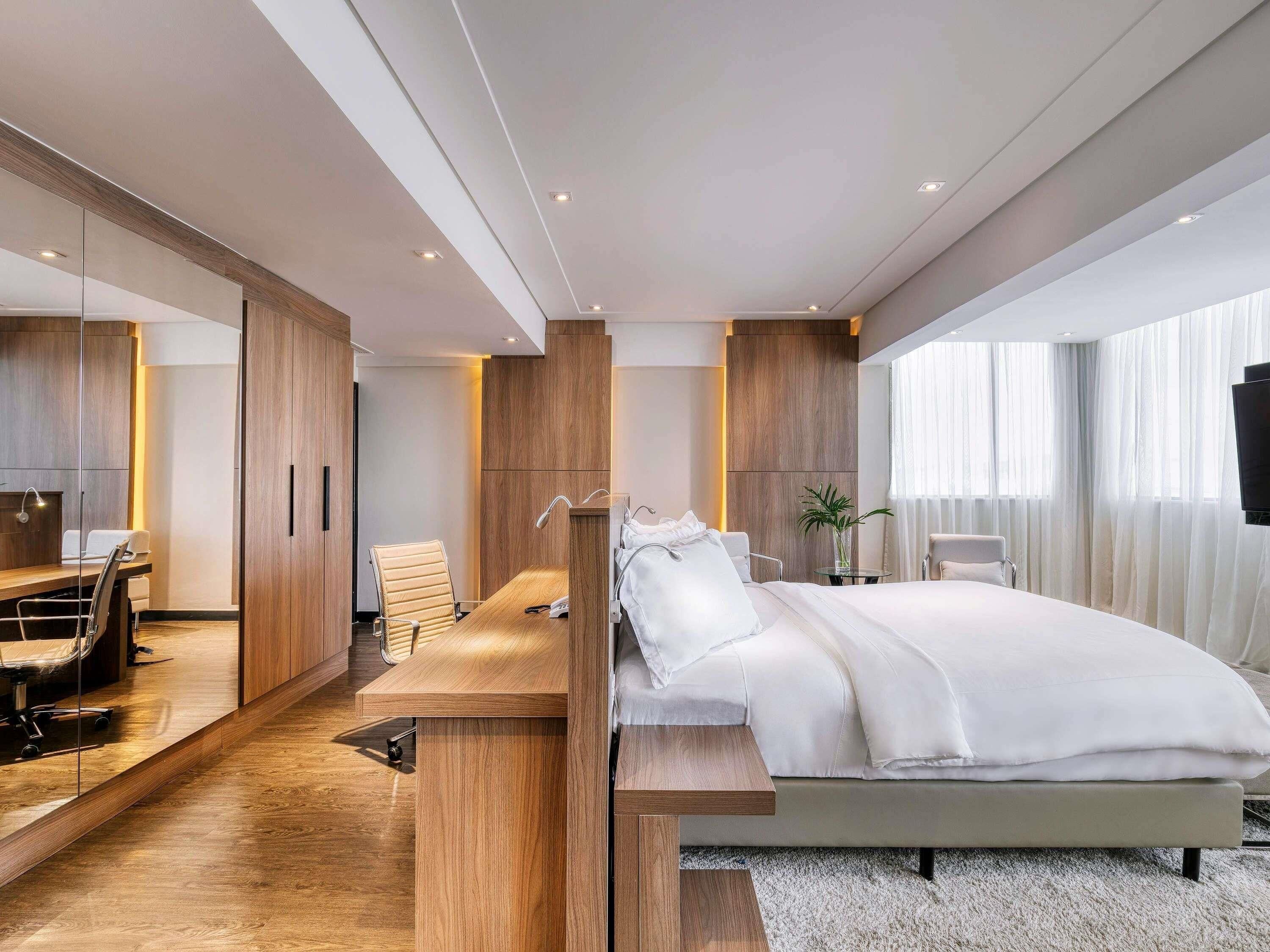 A modern hotel room with a large bed, a desk with a chair, a TV, and a large window.