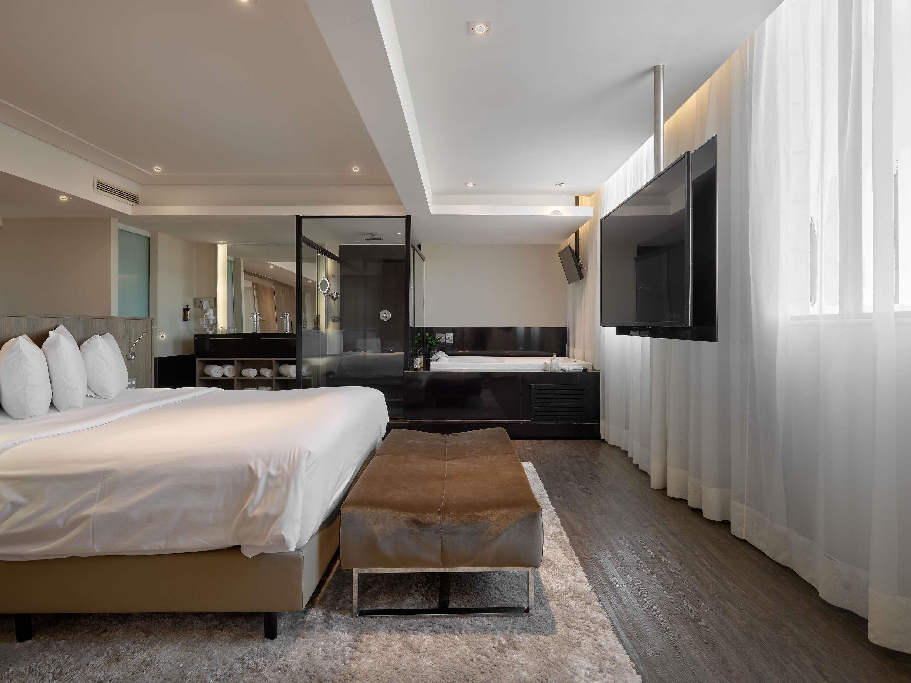 Una habitación de hotel moderna con una cama grande, una zona de estar, un baño con bañera y un televisor montado en la pared.