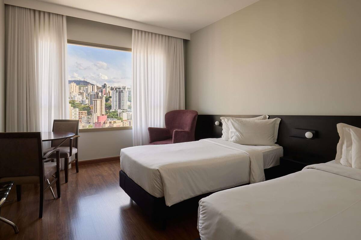 A hotel room with two beds, a desk, a chair, and a view of a cityscape.