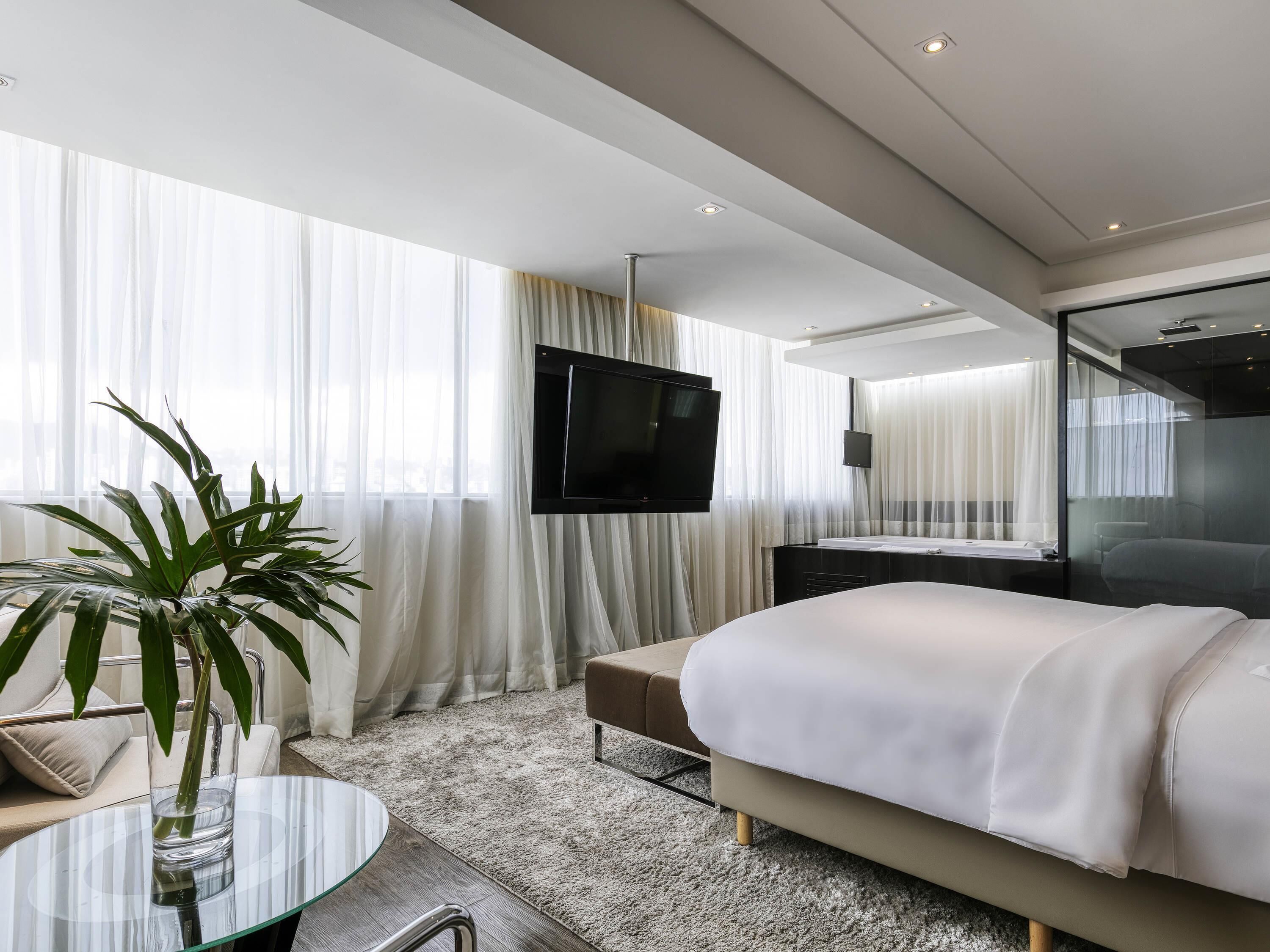 Quarto de hotel moderno com uma cama, uma TV, uma mesinha de vidro com uma planta e cortinas leves.
