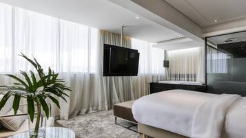 Quarto de hotel moderno com uma cama, uma TV, uma mesinha de vidro com uma planta e cortinas leves.