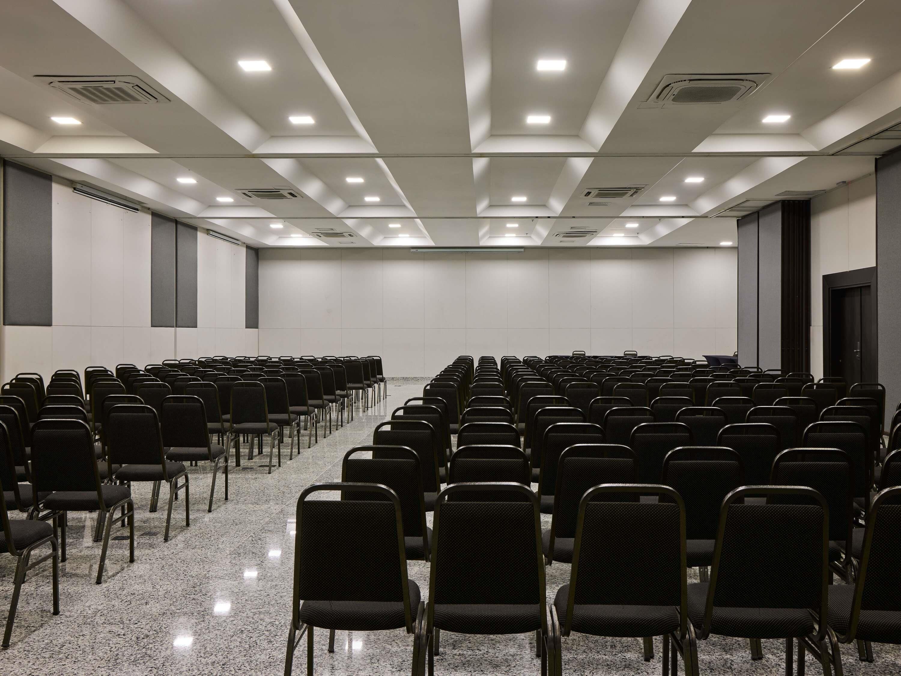 Un salón de conferencias con hileras de sillas, luces de techo y ventilación.