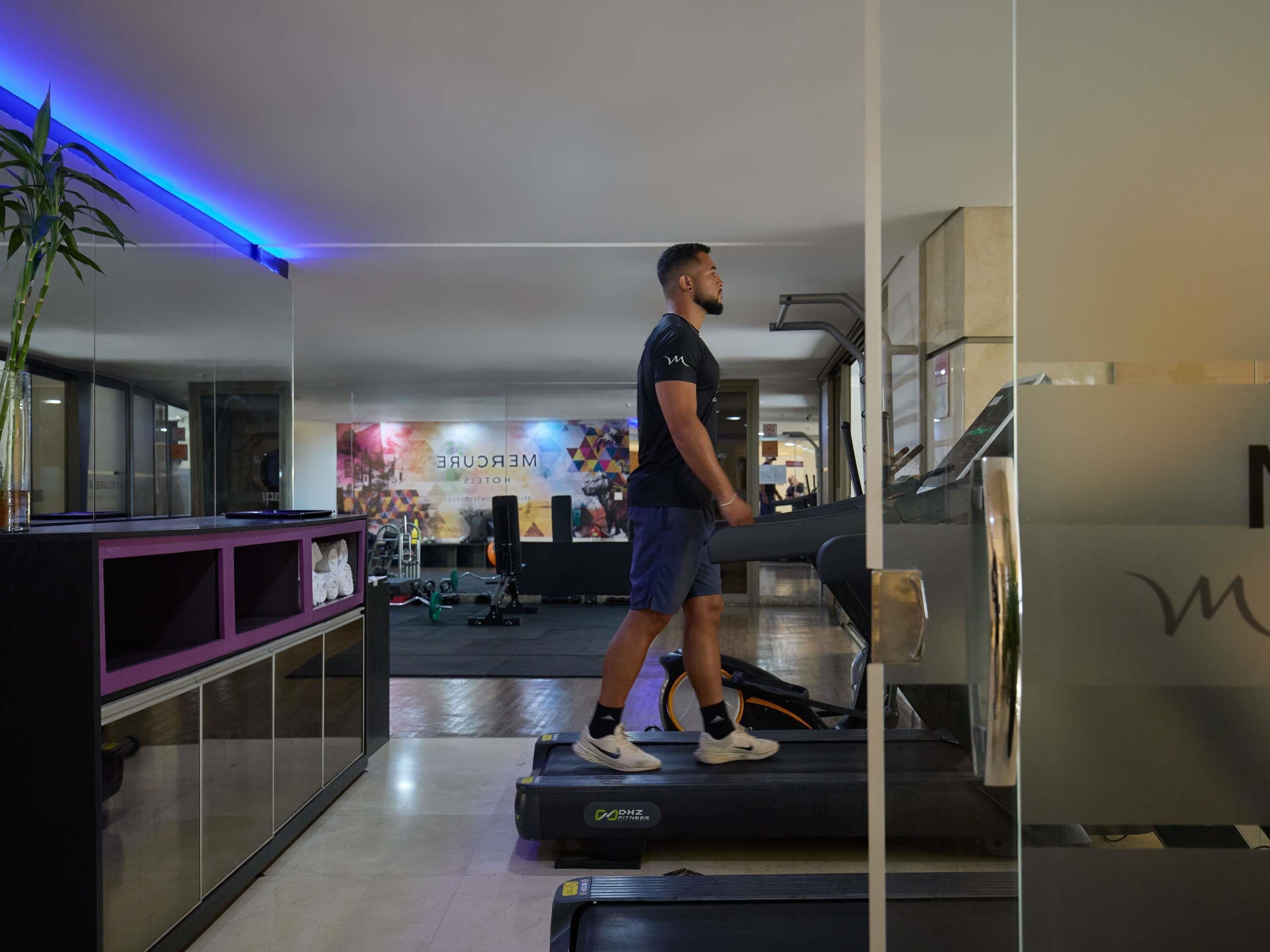 A man walking on a treadmill in a gym with mirrored walls, exercise equipment, and a colorful wall mural.