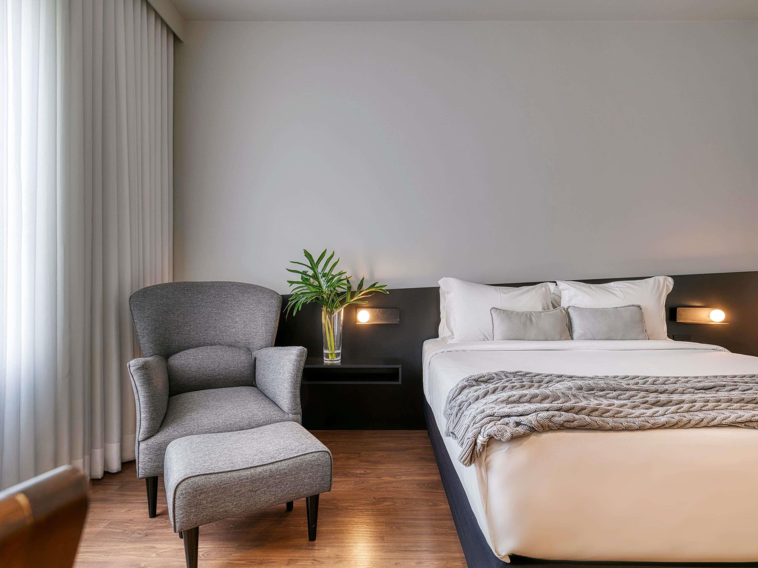 Quarto de hotel moderno com uma poltrona cinza, uma mesinha de cabeceira com uma planta, uma cama com roupa de cama branca e uma cabeceira escura.