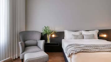 Quarto de hotel moderno com uma poltrona cinza, uma mesinha de cabeceira com uma planta, uma cama com roupa de cama branca e uma cabeceira escura.