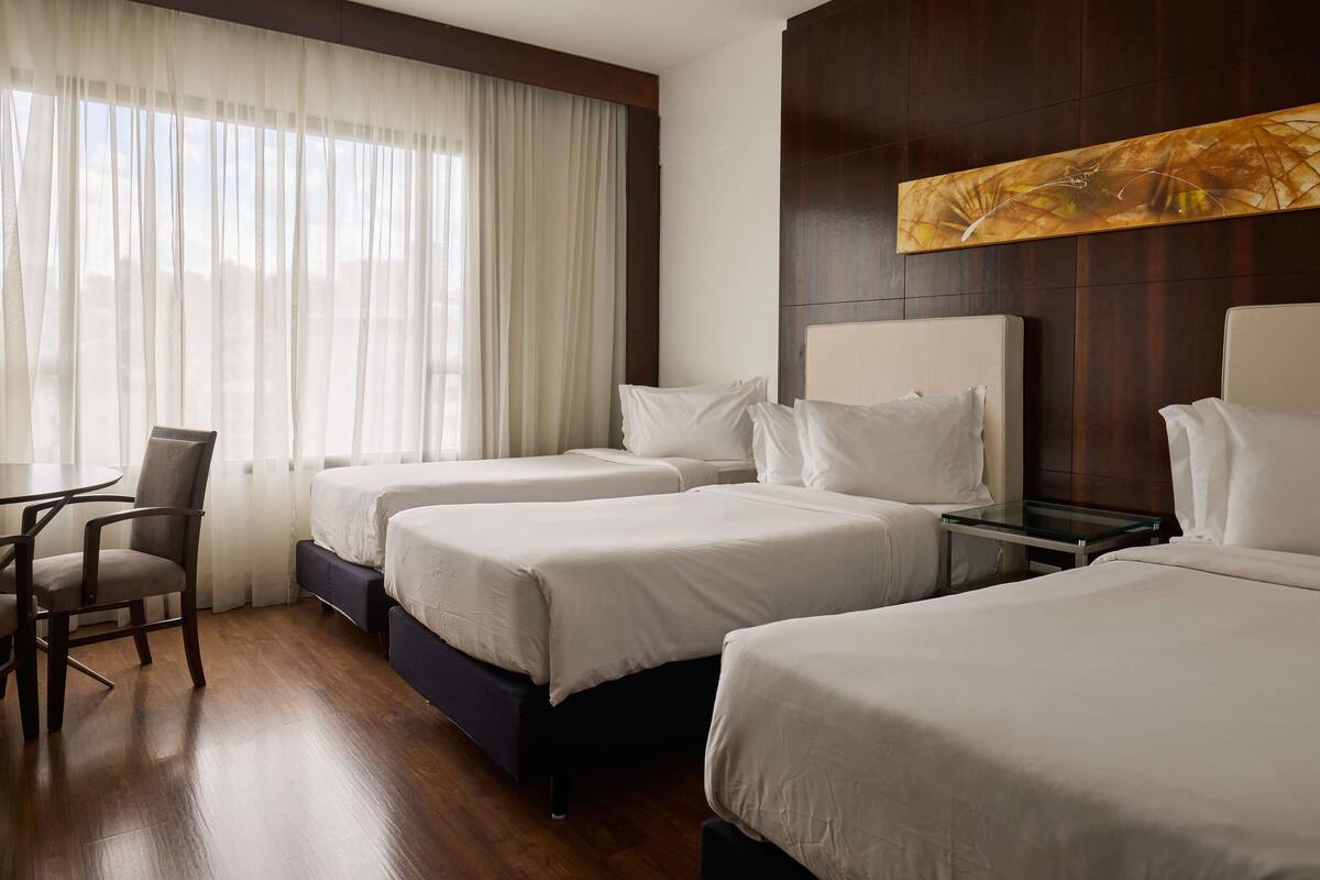 A hotel room with two beds, a chair, a small table, and a large window with sheer curtains.