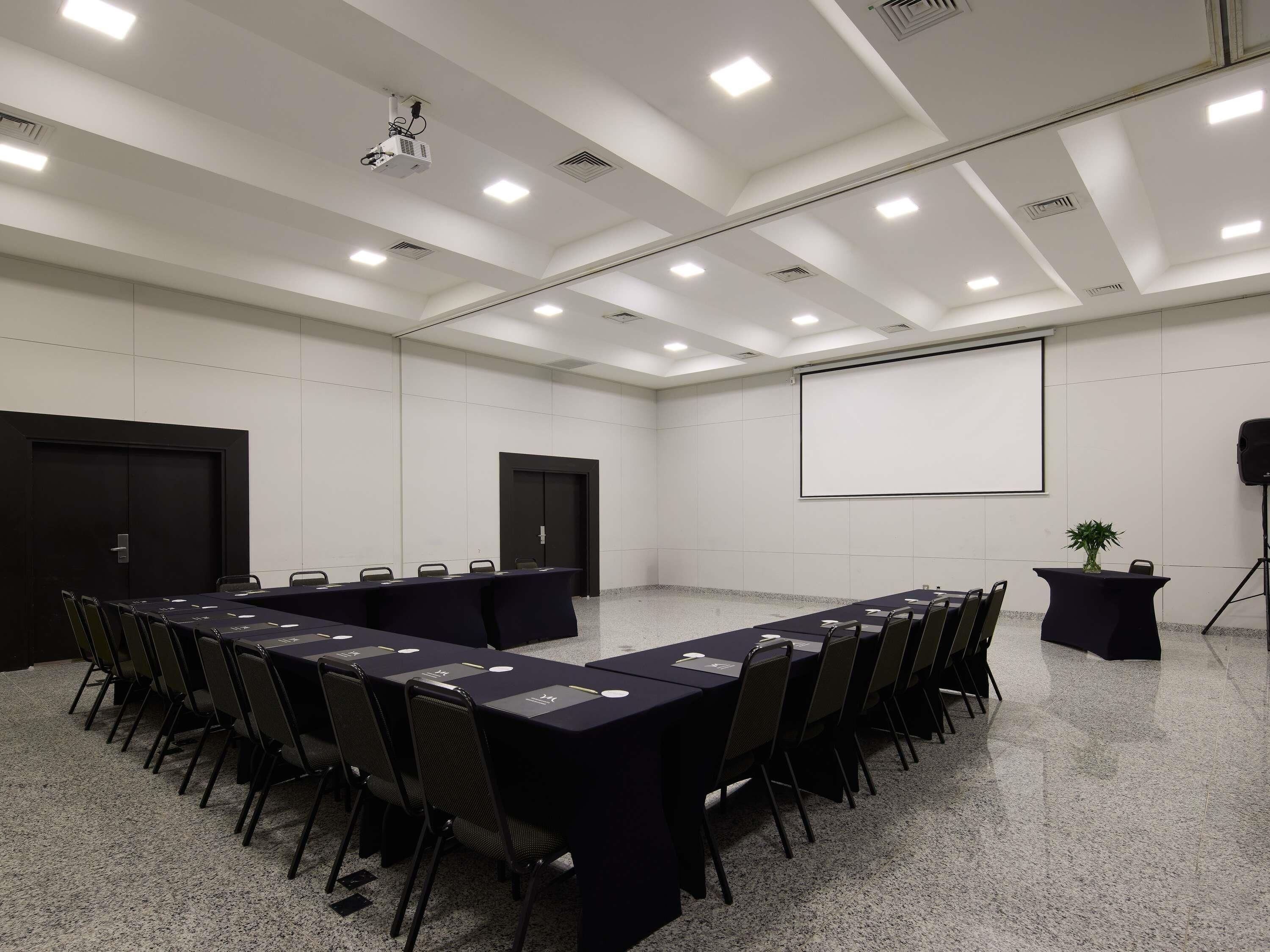 Un salón de conferencias con una larga mesa, sillas, un proyector, una pantalla y un orador.