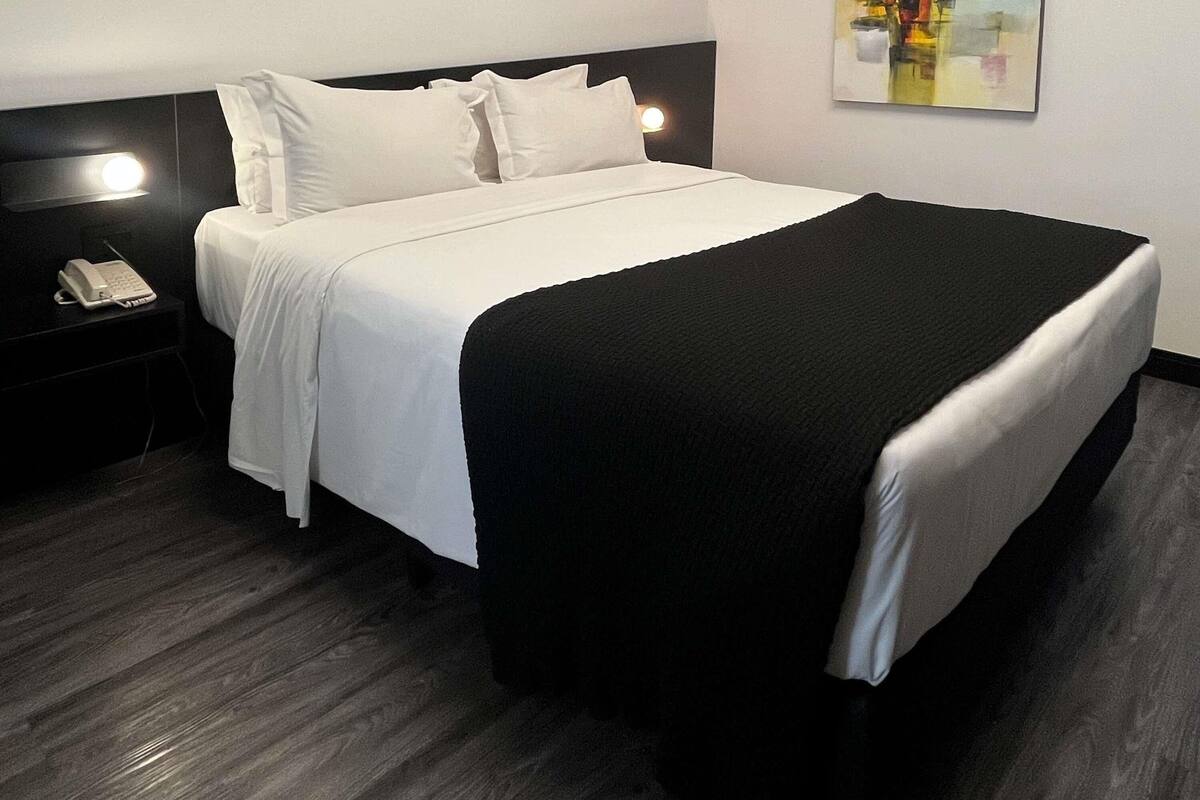 A hotel room with a bed, a nightstand with a phone, and a wall-mounted light.