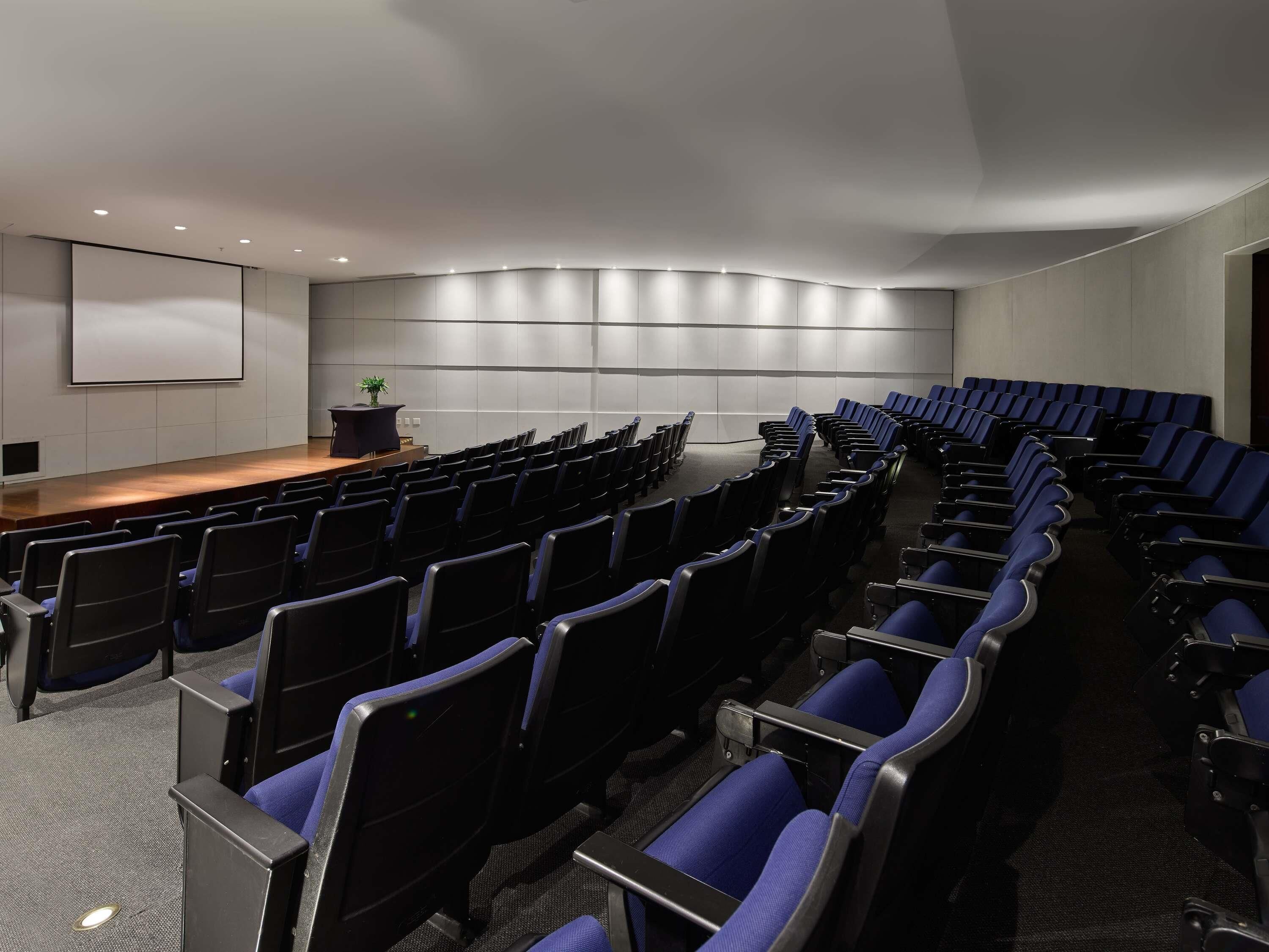 A conference room with rows of chairs, a stage with a screen, and a plant on the stage.
