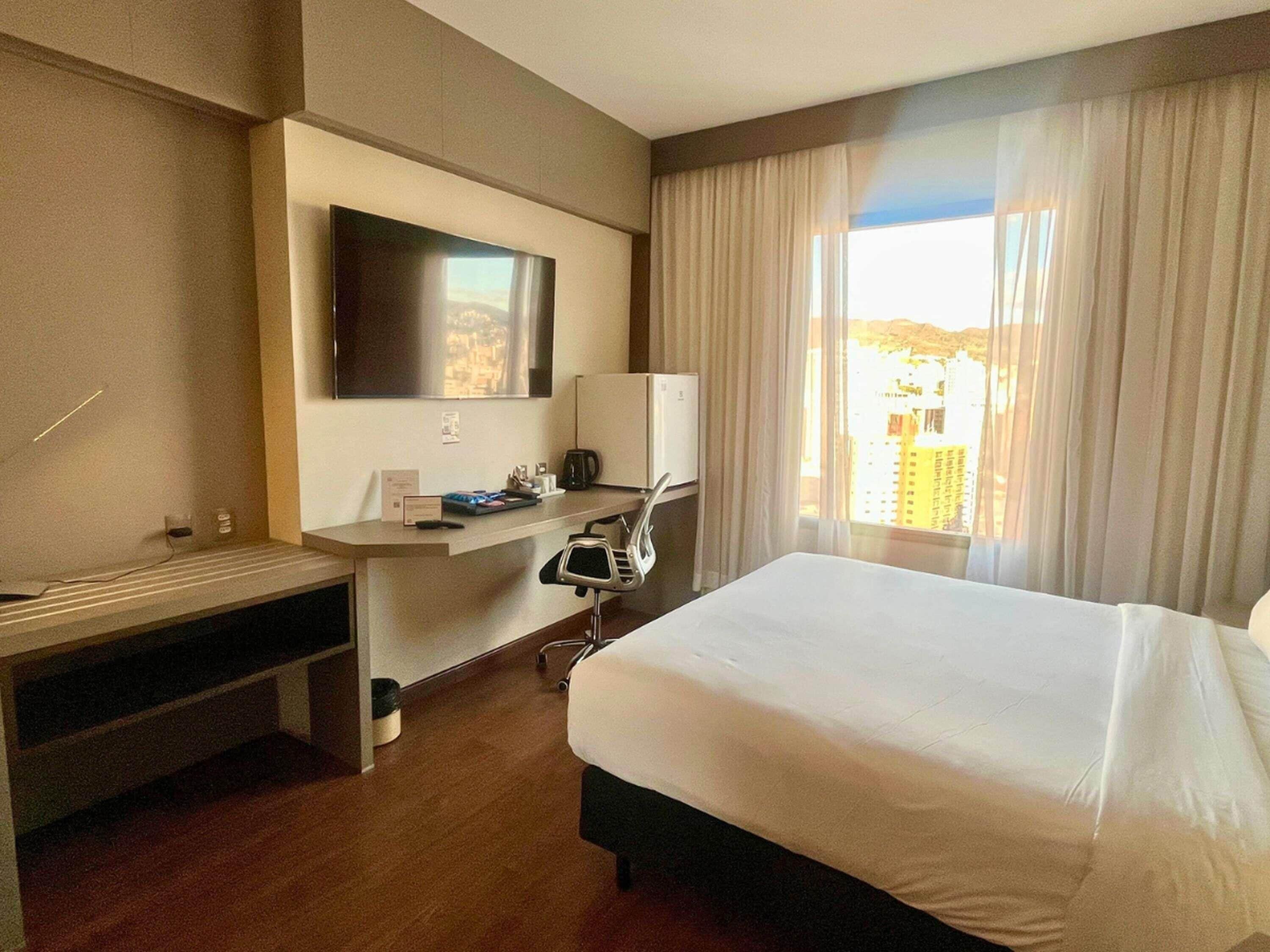 A hotel room with a bed, a desk with a flat-screen TV, a chair, and a window with curtains.