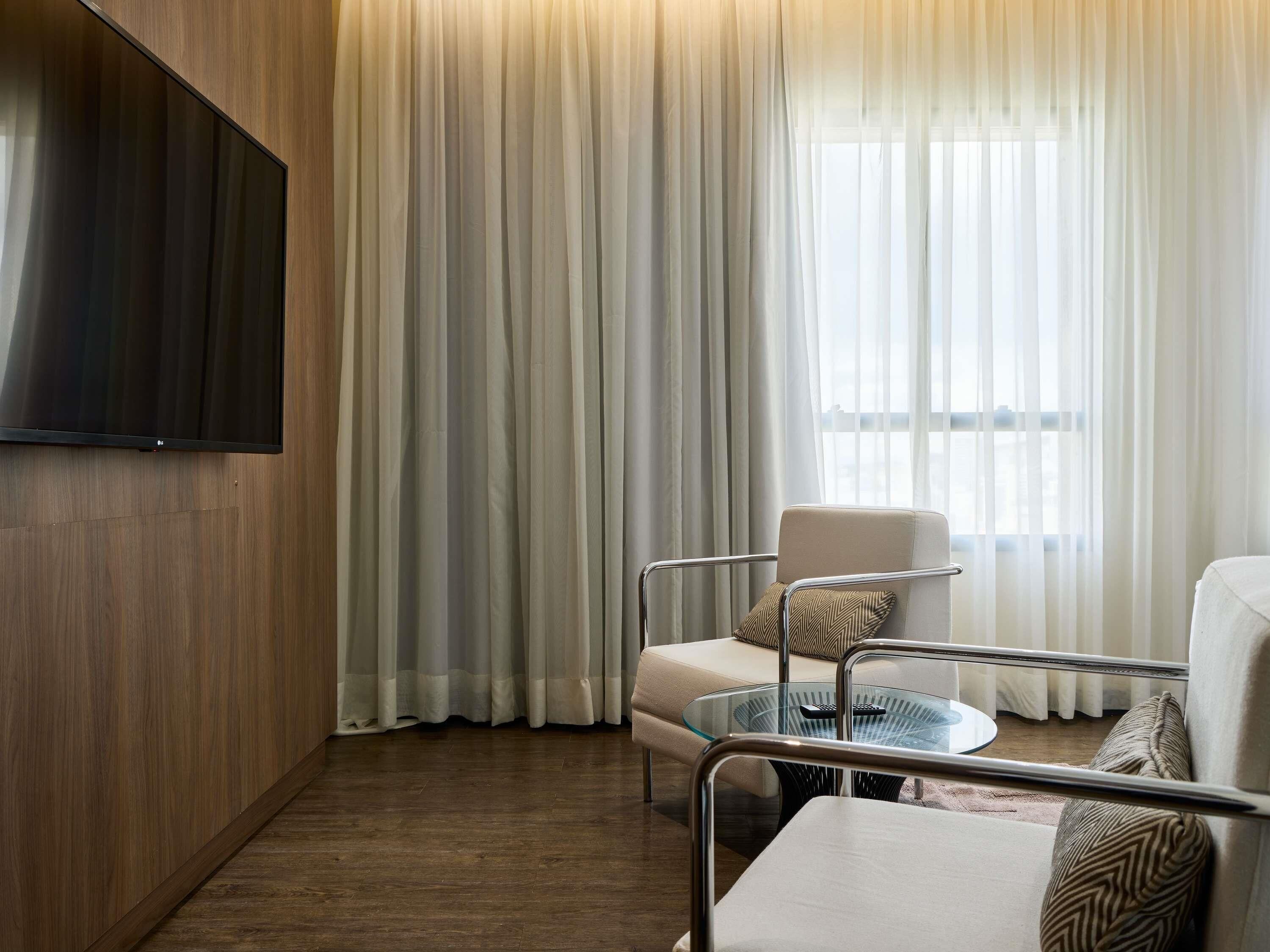 A modern hotel room with a flat-screen TV mounted on a wooden wall, a glass coffee table, a cushioned armchair, and a sofa with patterned cushions.