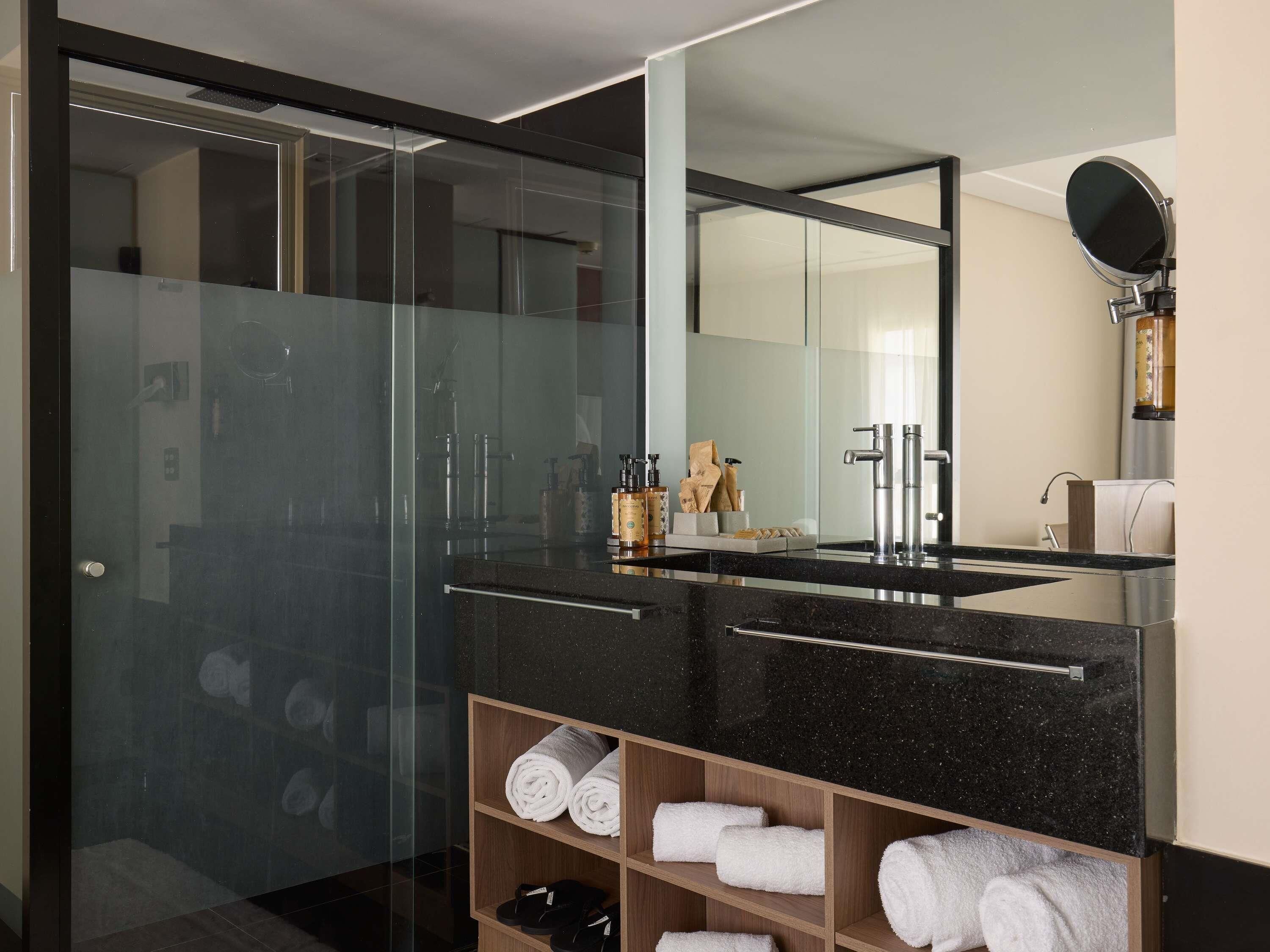 A modern bathroom with a glass shower enclosure, a black marble sink, and a wooden shelf with towels.