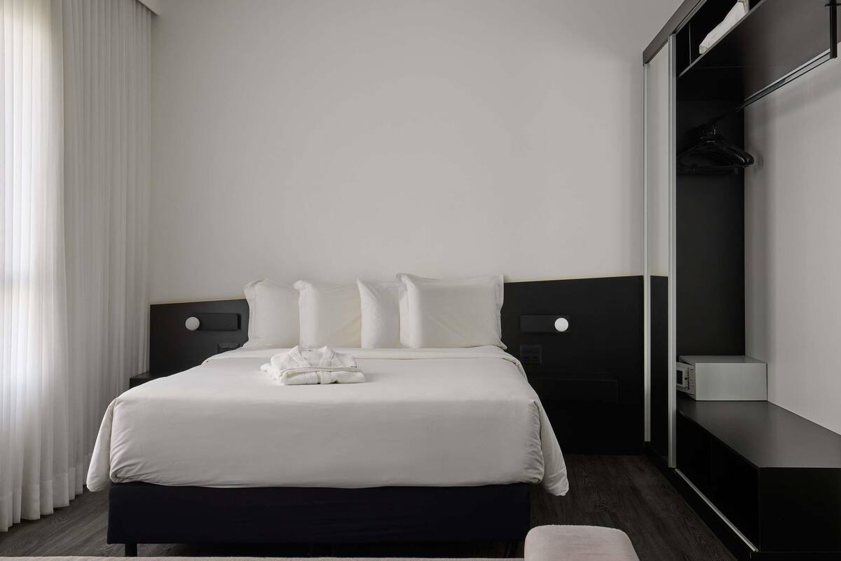 A modern hotel room with a neatly made bed, a dark headboard, a bedside table, and a wardrobe.