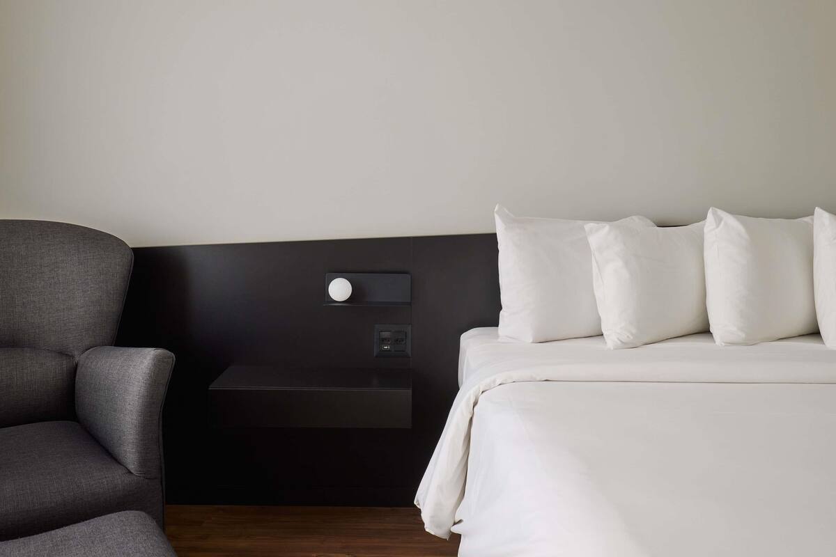 A hotel room with a grey armchair, a bed with white bedding, a black headboard, and a bedside table with a lamp.