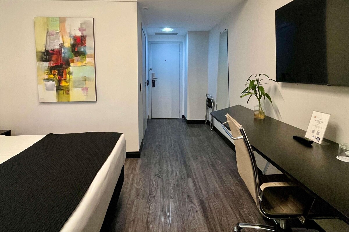 Hotellihuone, jossa on sänky, työpöytä tuolilla, televisio ja taulu seinällä.