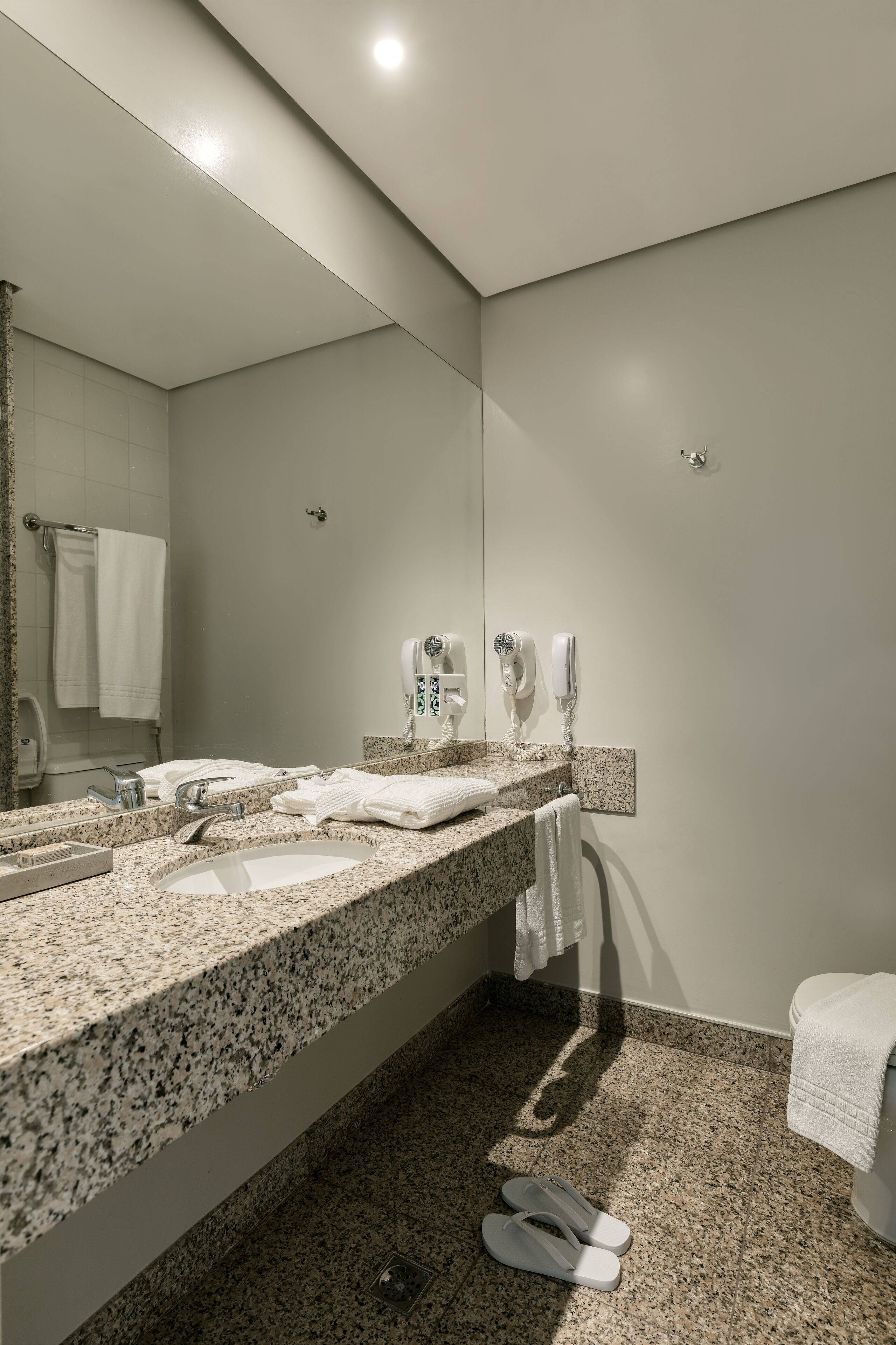 A modern bathroom with a granite countertop, a sink, a mirror, a towel rack, and a pair of slippers on the floor.