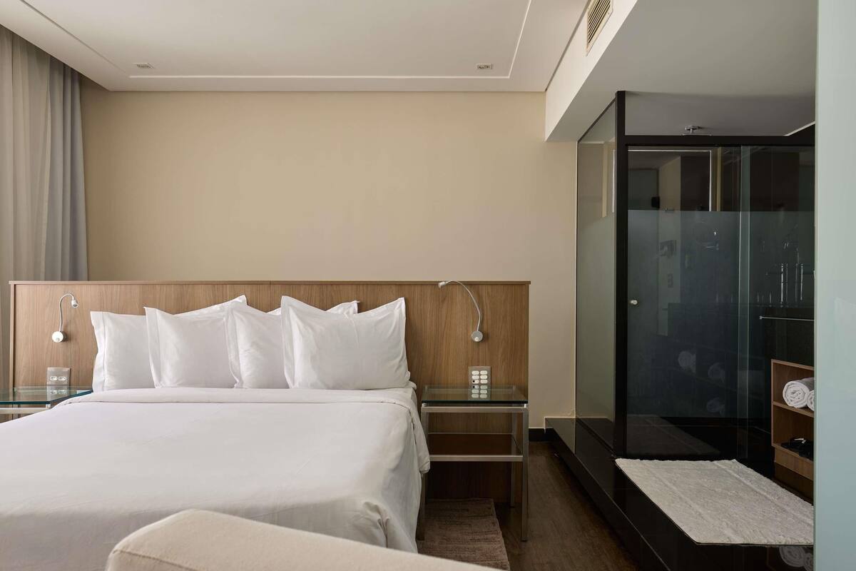 A hotel room with a bed, bedside tables, a control panel, a glass shower enclosure, and a wooden bench.