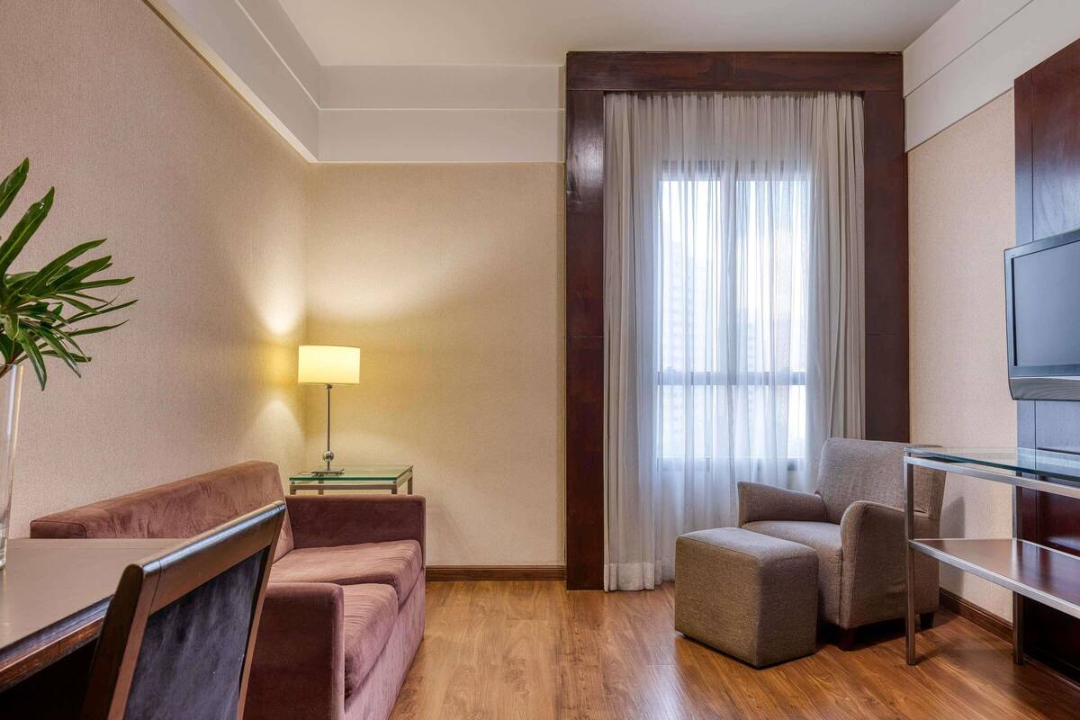 A hotel room with a wooden floor, a sofa, a chair, a small table, a lamp, a TV, and a window with curtains.