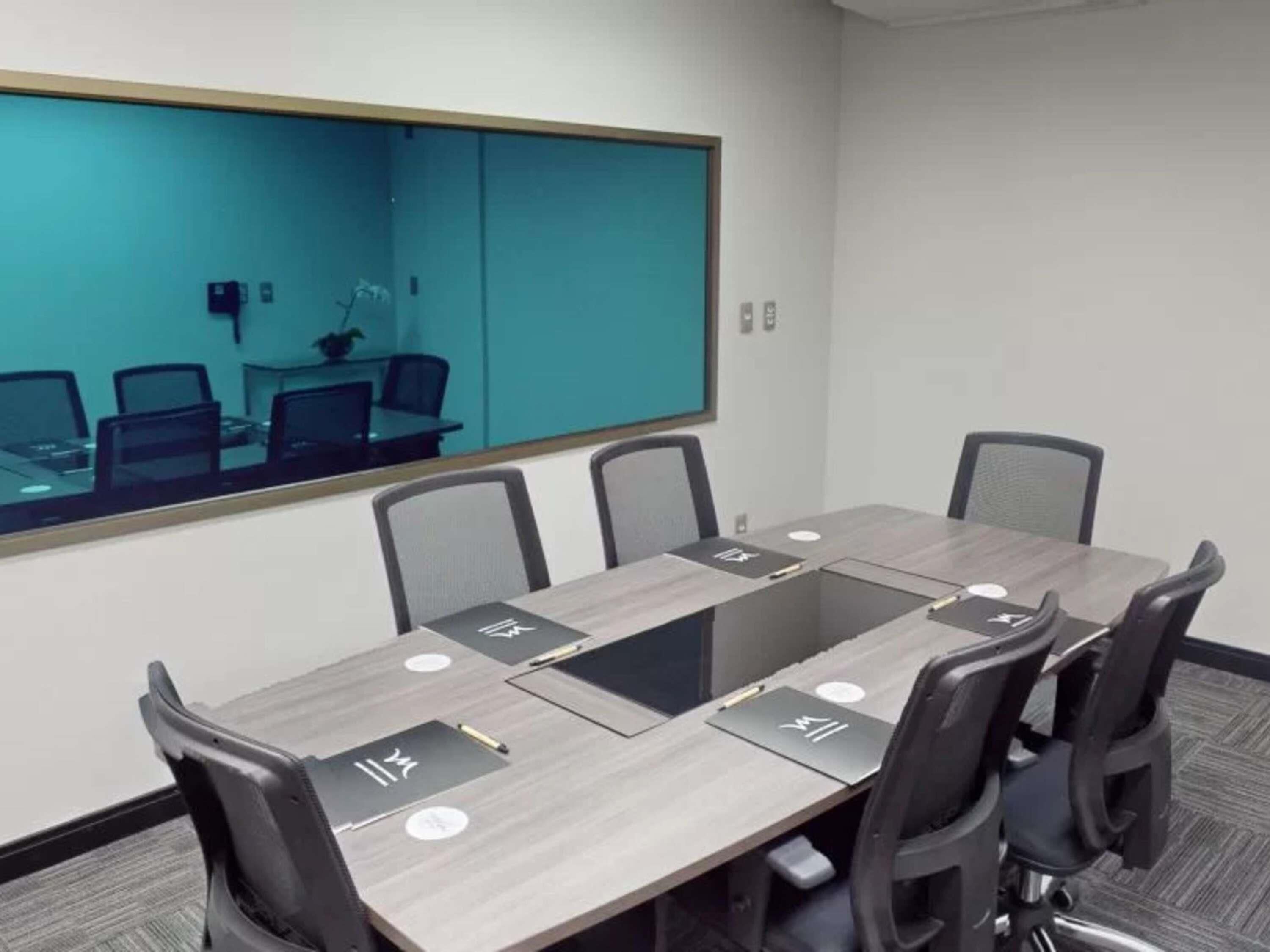 A conference room with a long table, six chairs, a large mirror, and a view of another room.
