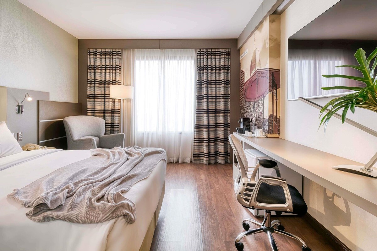 A hotel room with a bed, desk, chair, and a large plant.