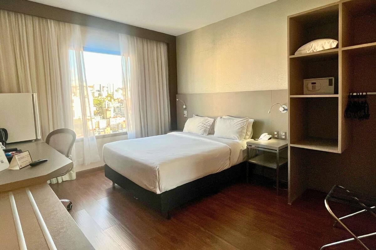 A hotel room with a bed, a desk, a chair, a nightstand, a lamp, a shelf with a pillow, and a view of the city through the window.