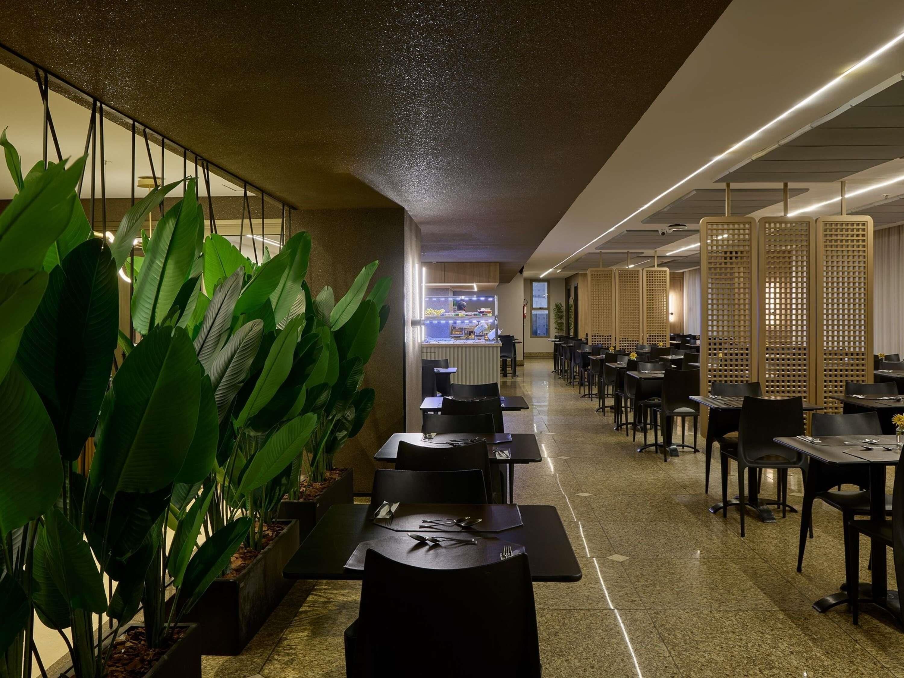 A modern restaurant with a long counter, black tables, and chairs. There are potted plants along the wall and a view of the kitchen in the background.