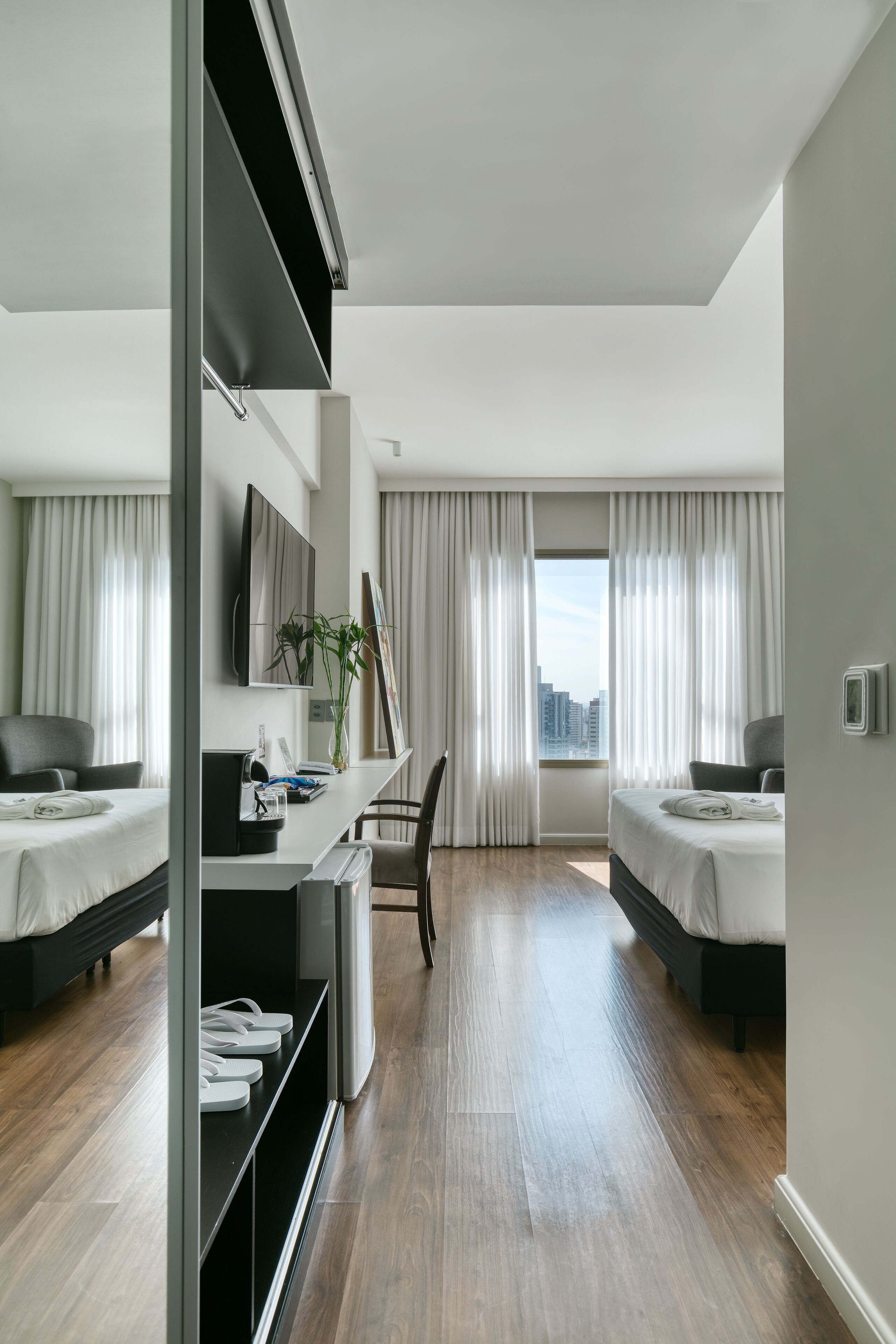 Une chambre d’hôtel moderne équipée de deux lits, d’un bureau avec un ordinateur, d’une chaise et offrant une vue sur la ville grâce à de grandes fenêtres.