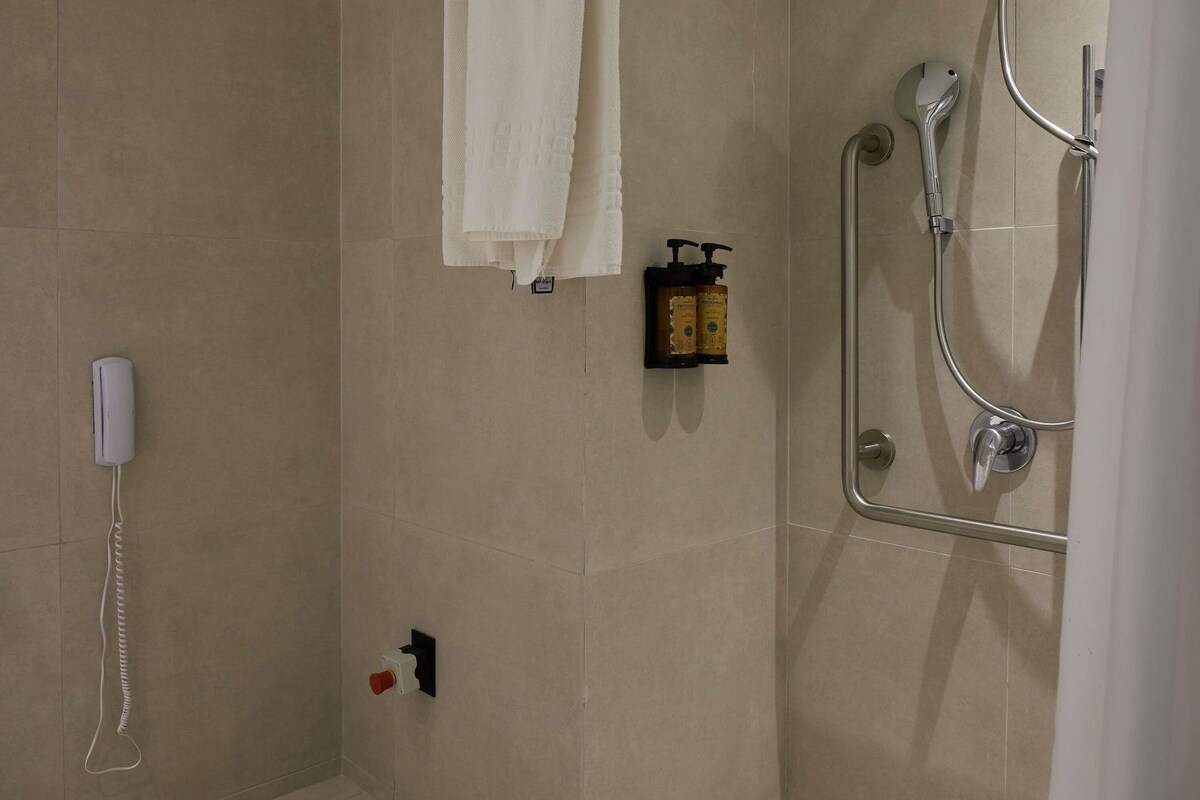 A bathroom with a shower, a towel hanging on the wall, a soap dispenser, and a phone mounted on the wall.