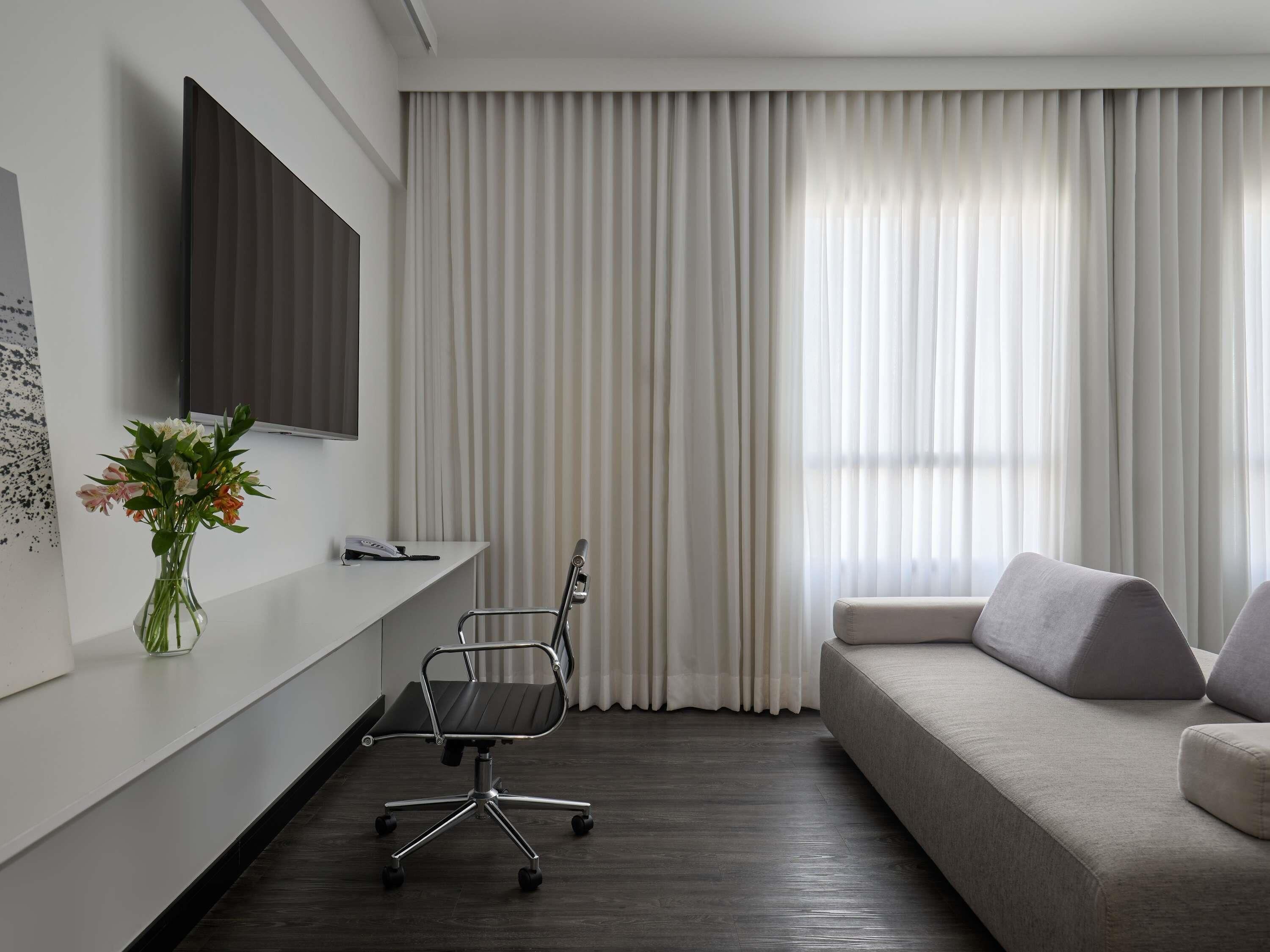Una habitación moderna con sofá, escritorio, silla, televisor en la pared y ventana con cortinas translúcidas.