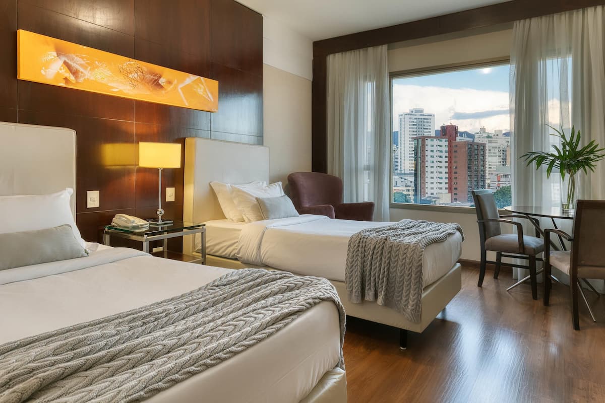 A hotel room with two beds, a desk, a chair, a table, and a large window with a city view.