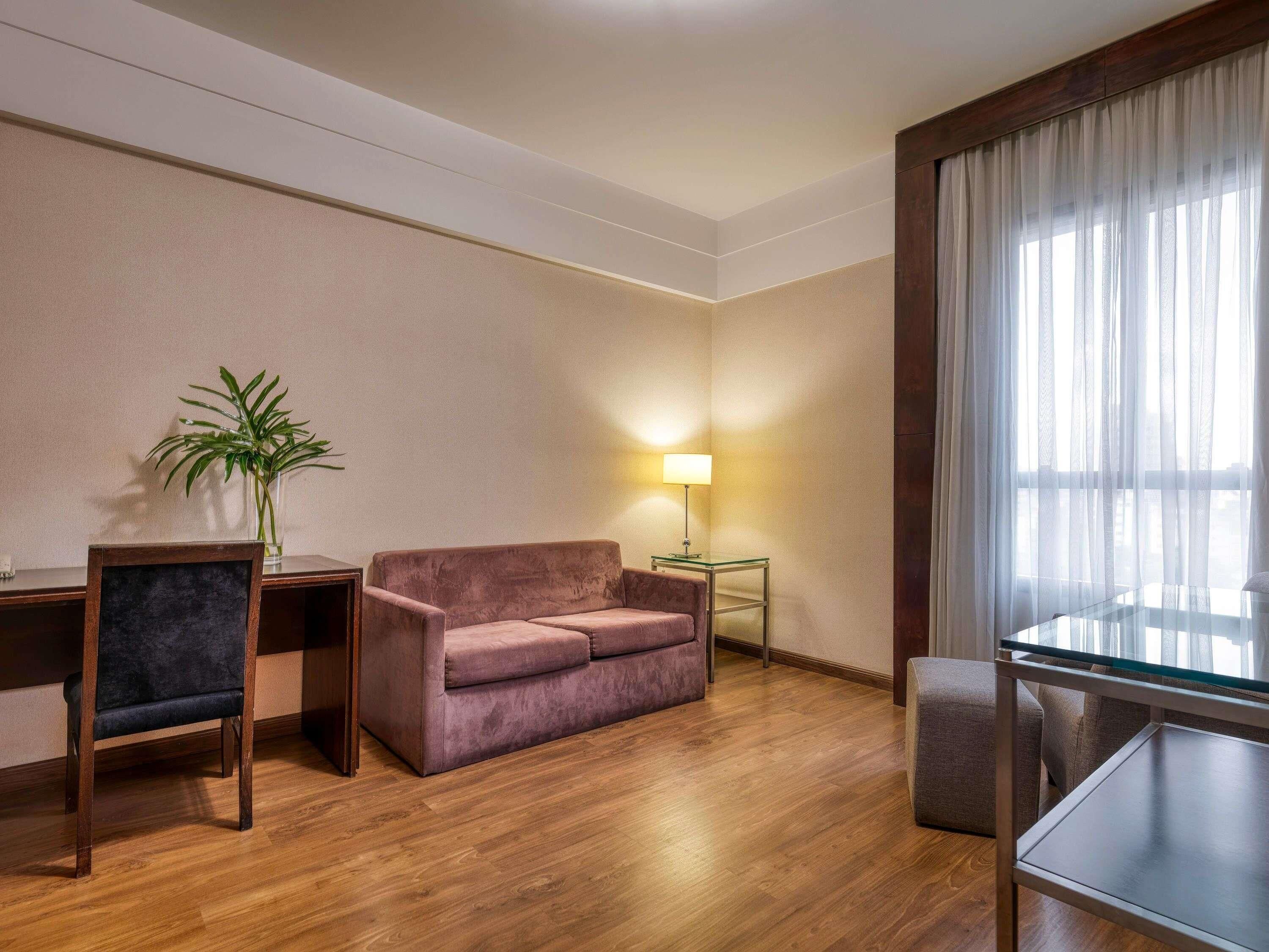 A hotel room with a wooden floor, a sofa, a desk with a chair, a small table with a lamp, and a large window with sheer curtains.