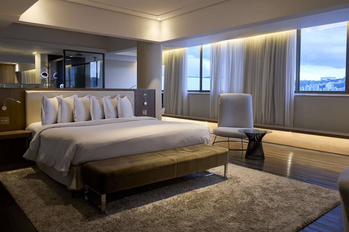 A modern hotel room with a large bed, a bench, a chair, a small table, and a view of the city through large windows.