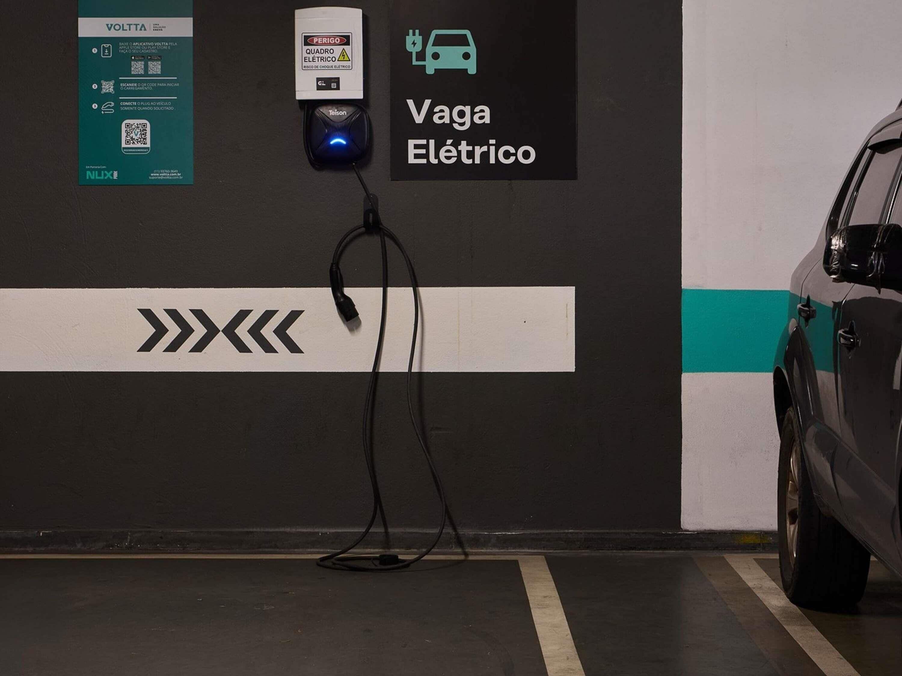 Una estación de carga para vehículos eléctricos con un código QR, un cartel que dice 'Vaga Elétrico' y un coche estacionado cerca.